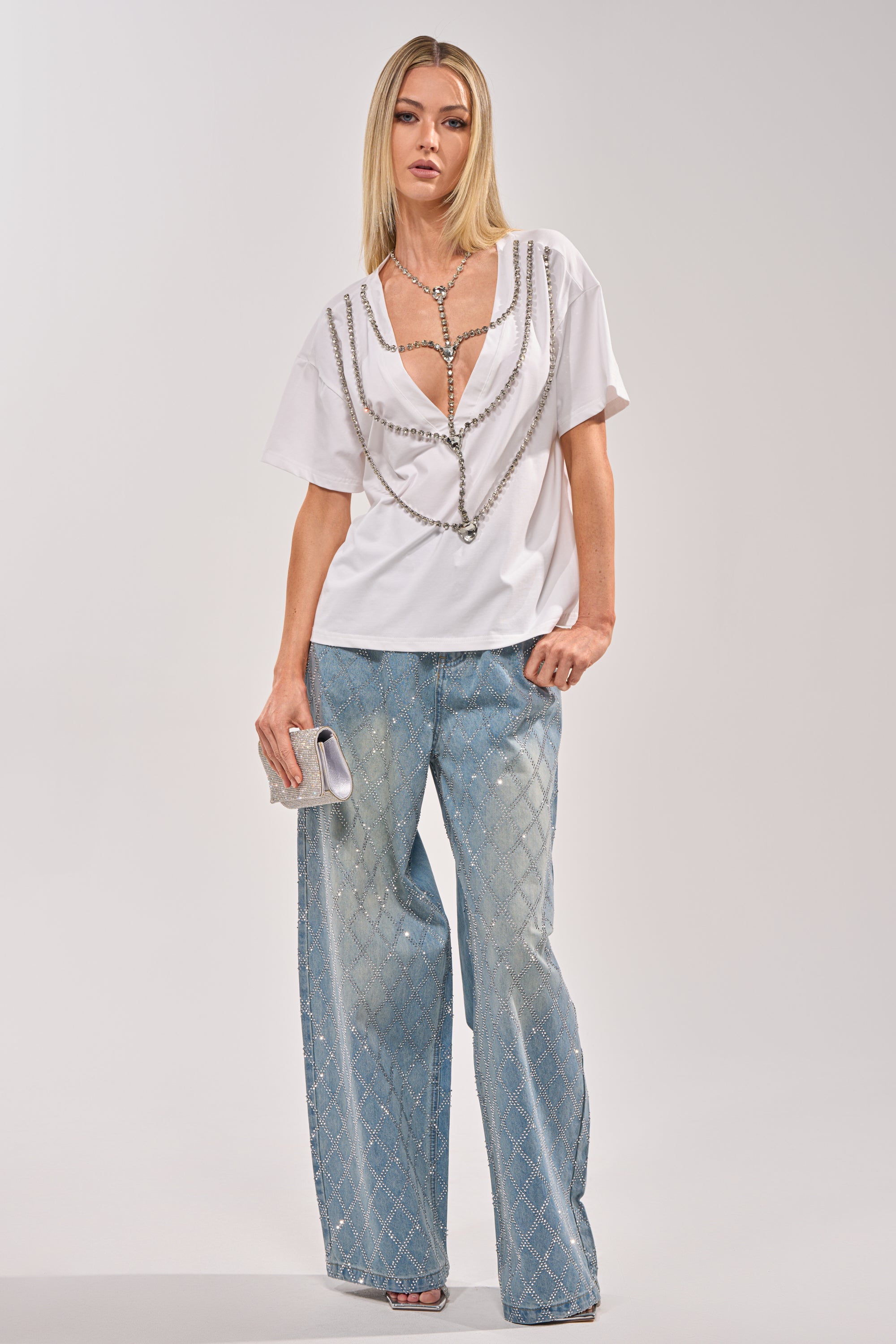 A woman with straight blonde hair and neutral makeup stands against a plain background wearing the DIAMONDS DANCING RHINESTONE DETAIL TEE, light blue wide-leg diamond pattern pants, and holding a small silver clutch. 7