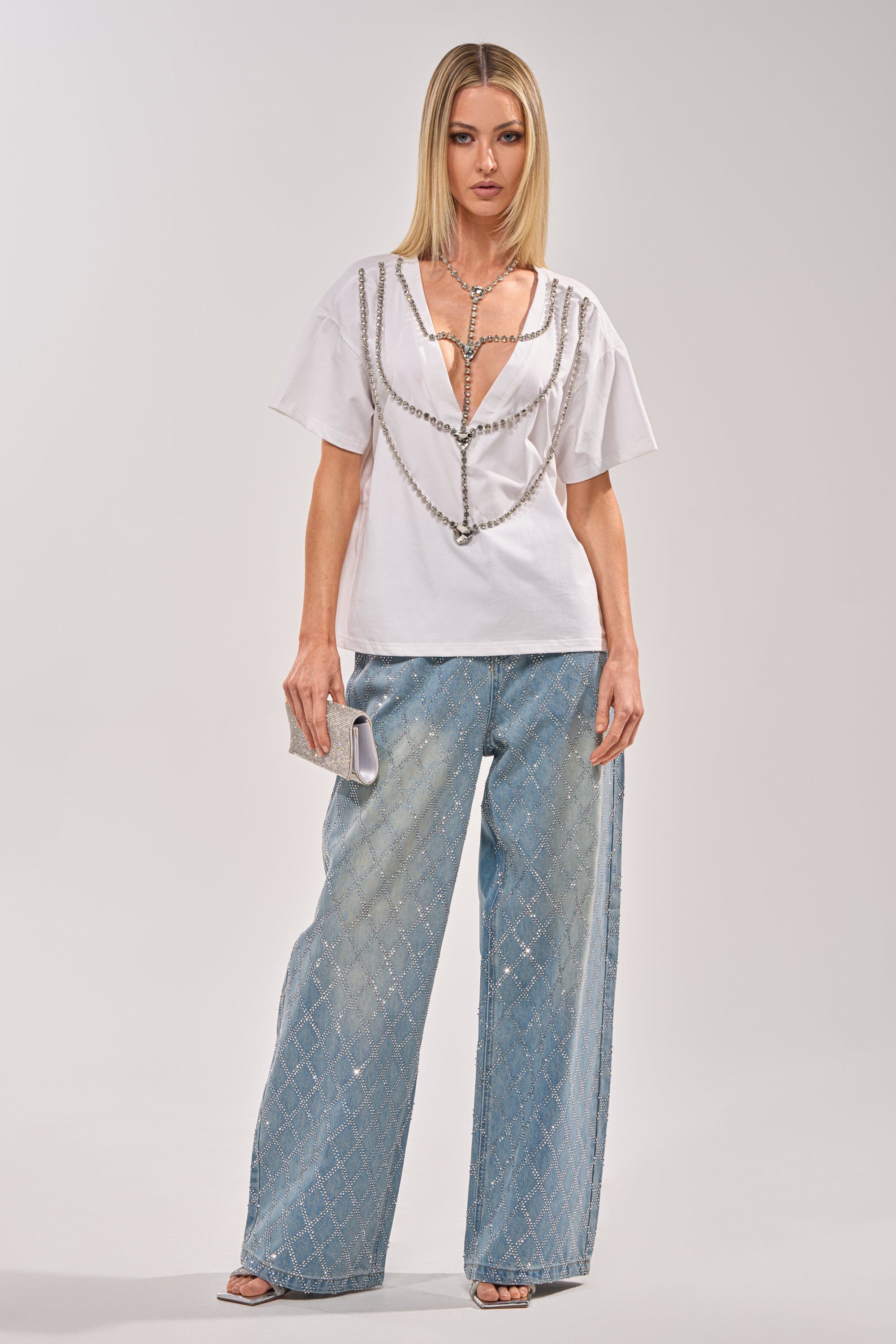 A woman with straight blonde hair wears the DIAMONDS DANCING RHINESTONE DETAIL TEE, light blue wide-leg diamond-patterned jeans, silver heels, and a silver clutch bag while standing against a plain background. 8
