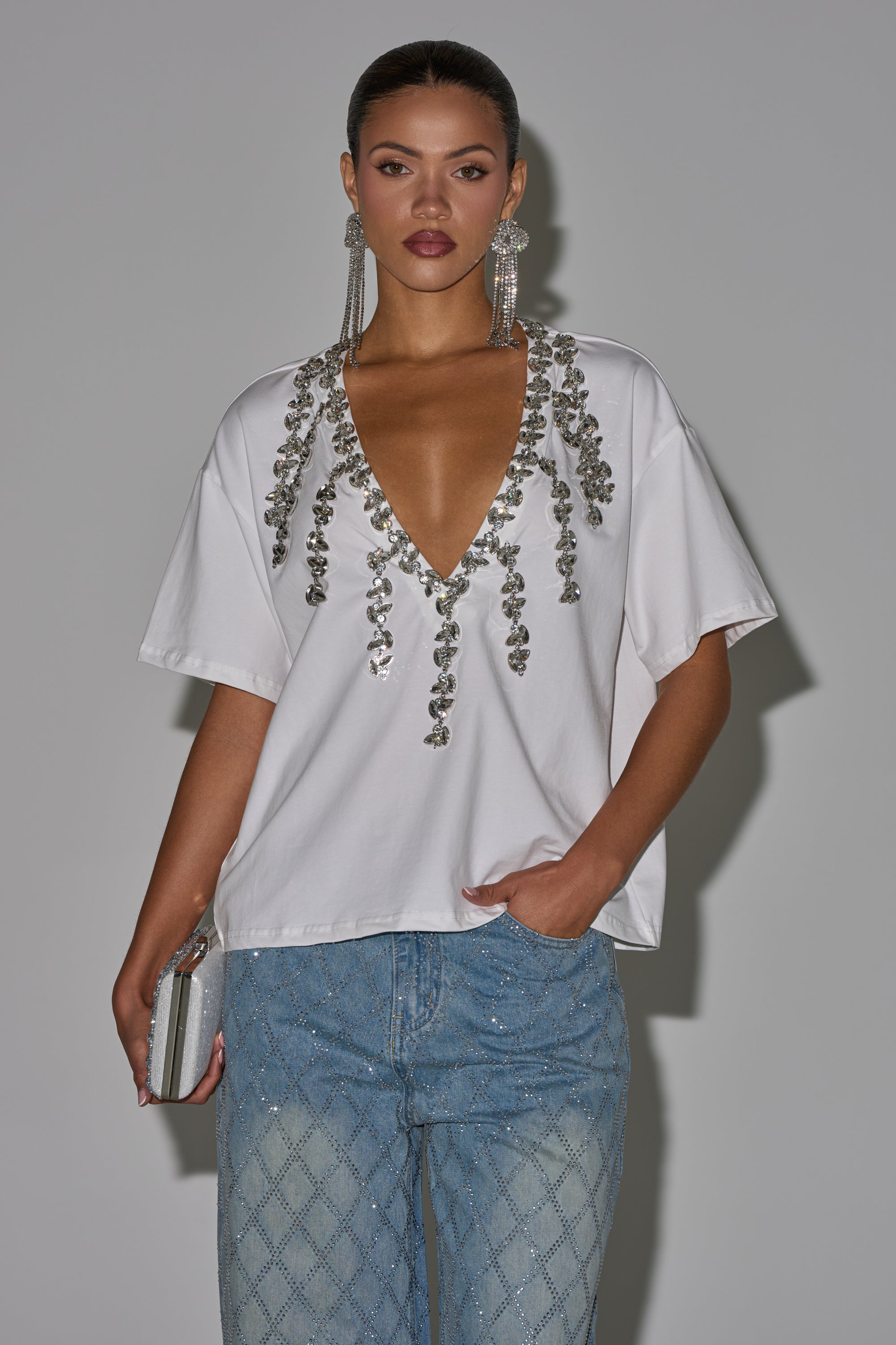 A woman poses against a gray background in the TOO DEEP RHINESTONE TEE, light blue jeweled jeans, long rhinestone earrings, and holding a silver clutch. 0