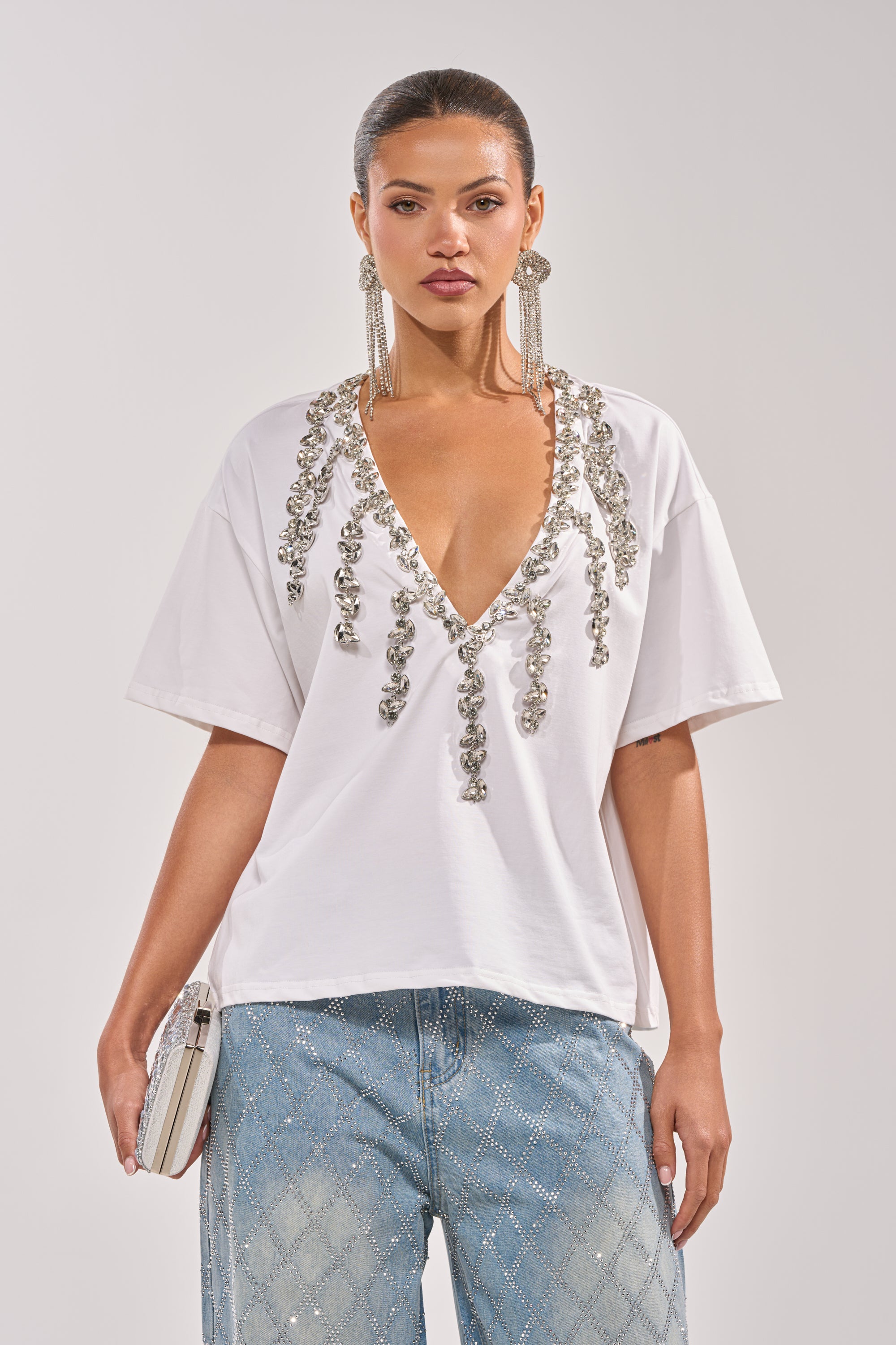 A woman wears the TOO DEEP RHINESTONE TEE with sparkling embellishments, blue jeans with a diamond pattern, long silver earrings, and holds a small white clutch against a plain light background. 1