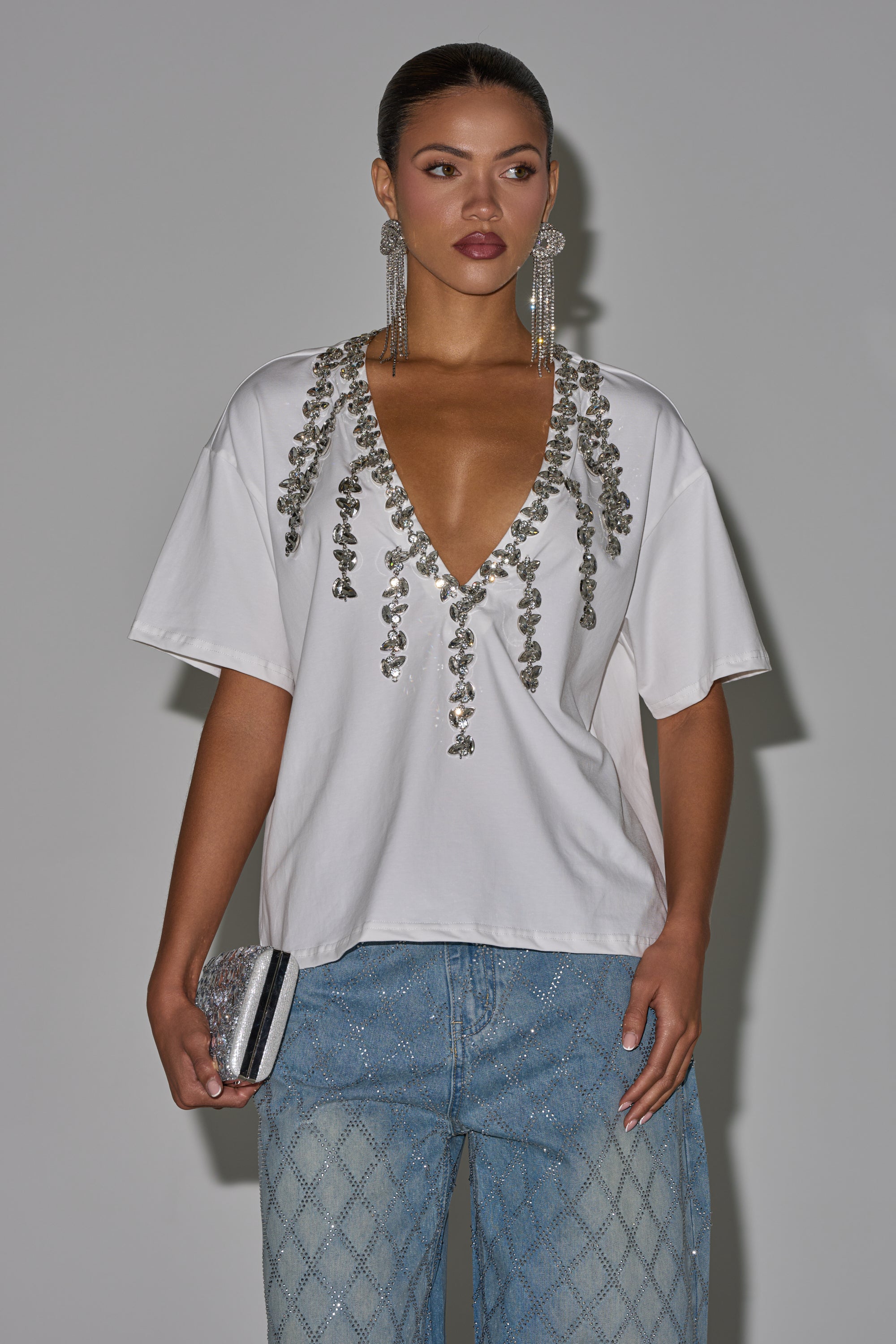 A woman poses against a plain background wearing the TOO DEEP RHINESTONE TEE with silver accents, blue patterned jeans, long dangling earrings, and holding a silver clutch. 2