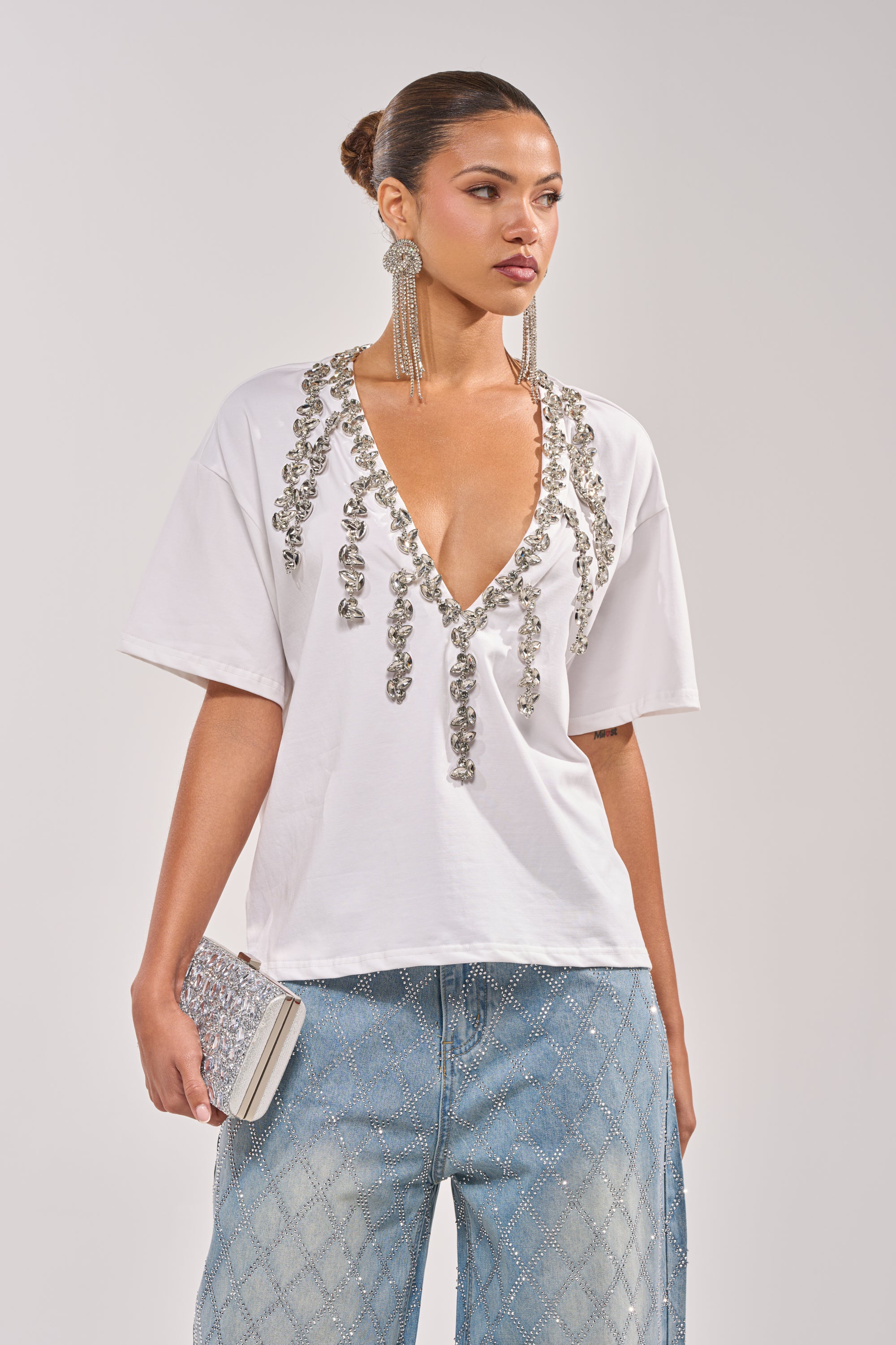 A woman wears the TOO DEEP RHINESTONE TEE, a V-neck shirt with silver embellishments, paired with wide-leg light blue diamond-patterned jeans, large dangling earrings, and a sparkling silver clutch. 3