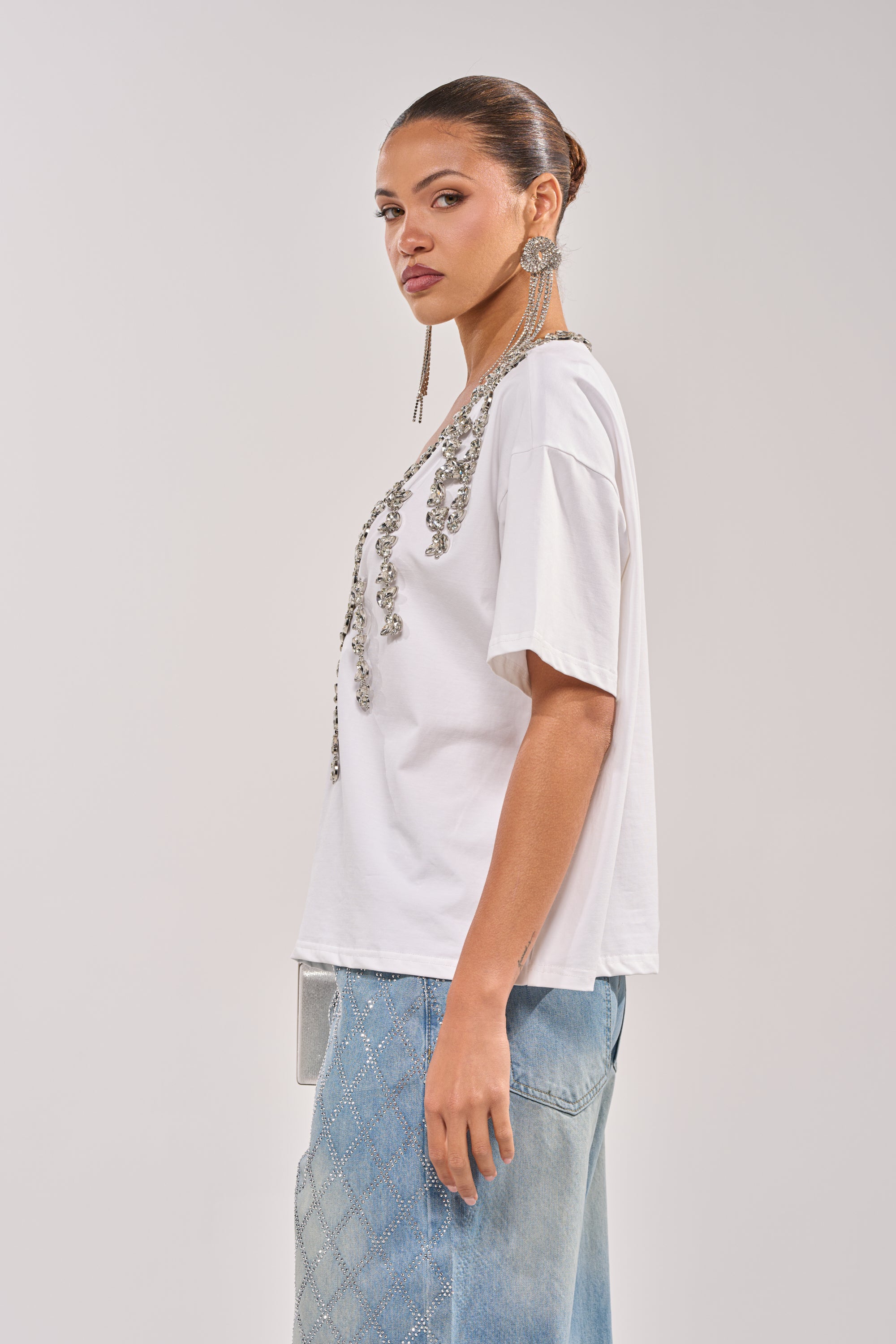 A woman with her hair in a bun wears the TOO DEEP RHINESTONE TEE, light blue patterned jeans, and statement earrings. She stands sideways against a plain, light background. 4