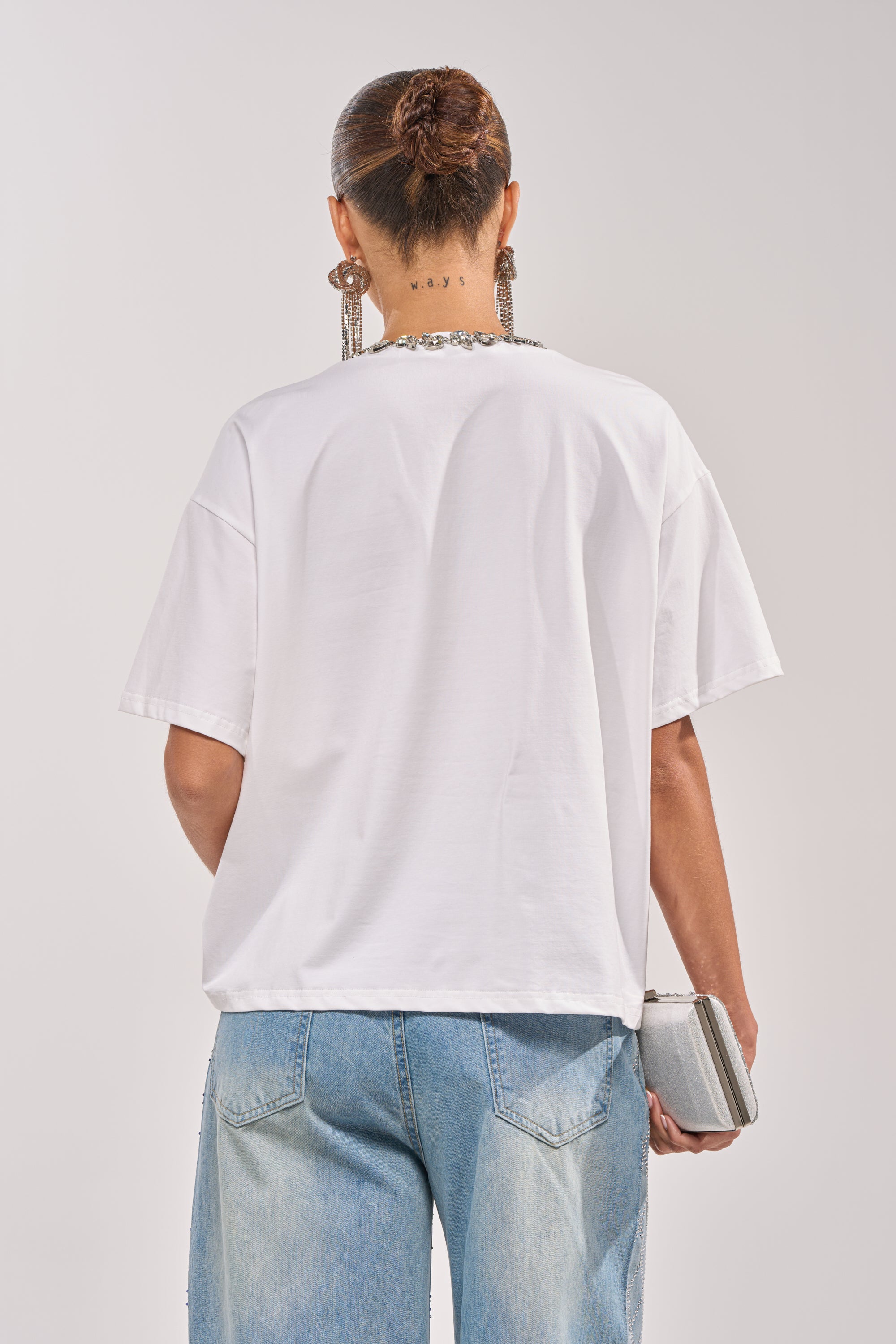 A person with hair in a bun, wearing large earrings and the TOO DEEP RHINESTONE TEE, paired with light blue jeans, stands facing away and holds a small silver clutch in their right hand. 5