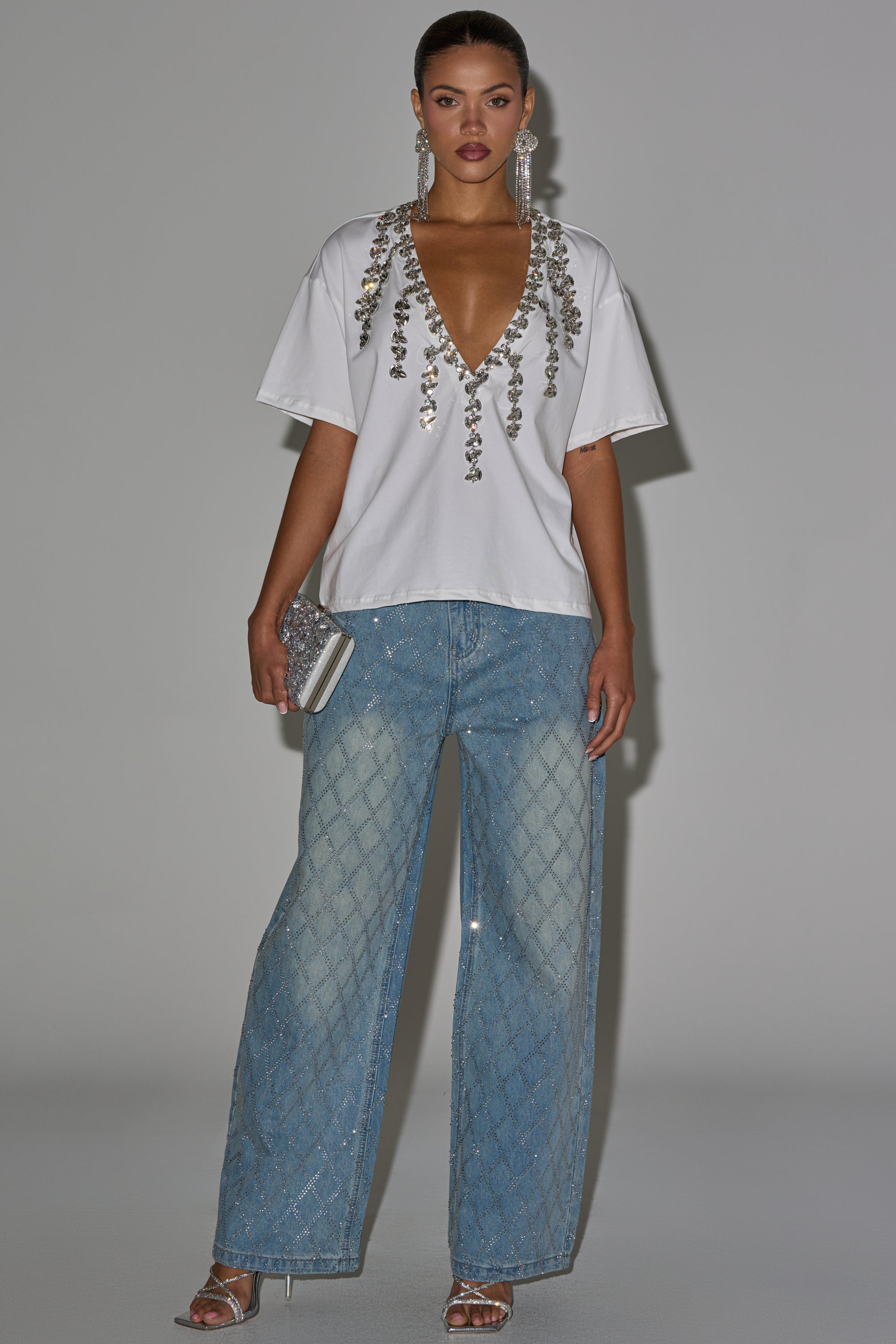 A woman stands against a plain background wearing the TOO DEEP RHINESTONE TEE, loose light blue checkered jeans, silver high-heeled sandals, and statement earrings. 6