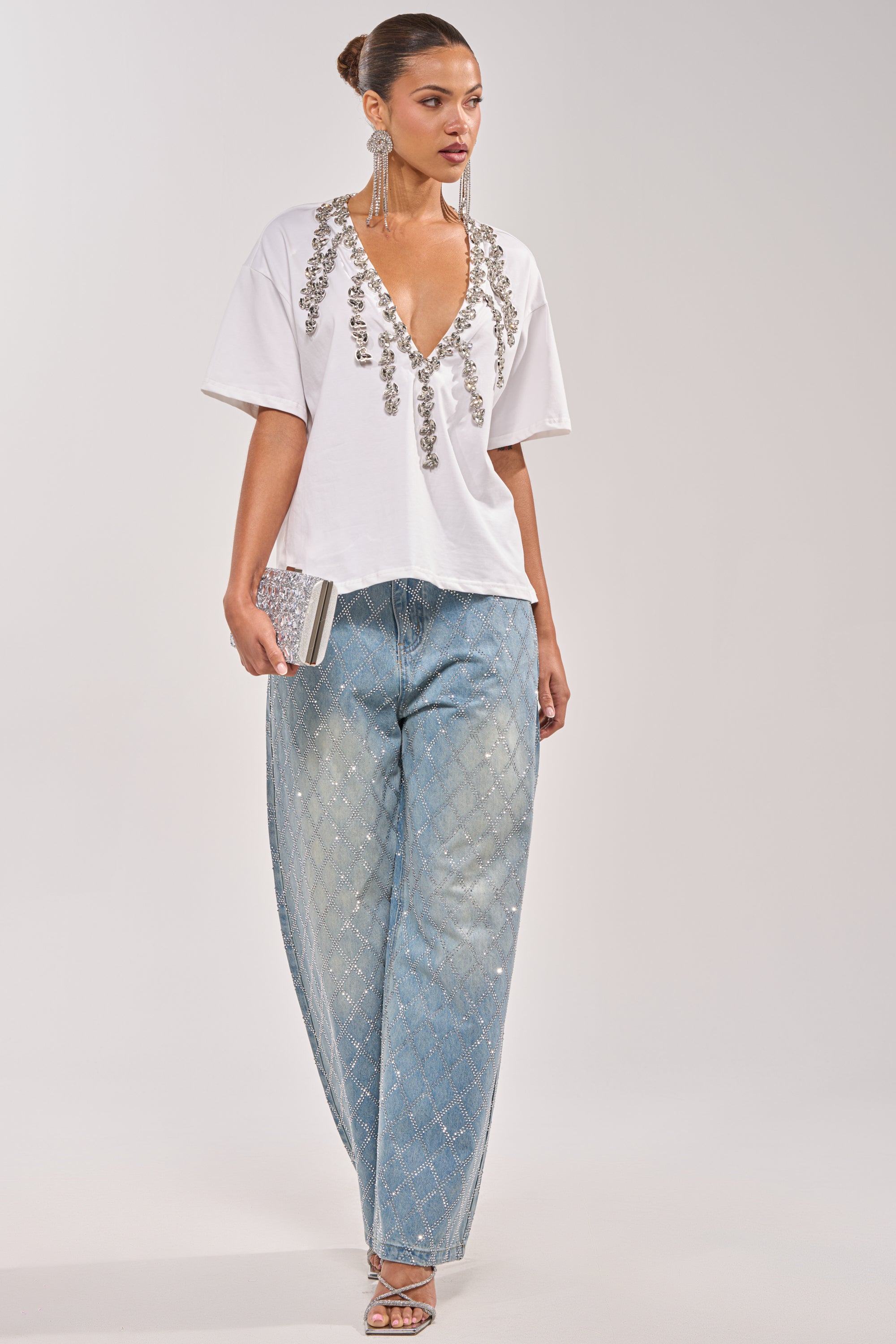 A woman wearing the TOO DEEP RHINESTONE TEE, wide-leg light blue diamond-pattern jeans, silver strappy heels, statement earrings, and holding a silver clutch. 7