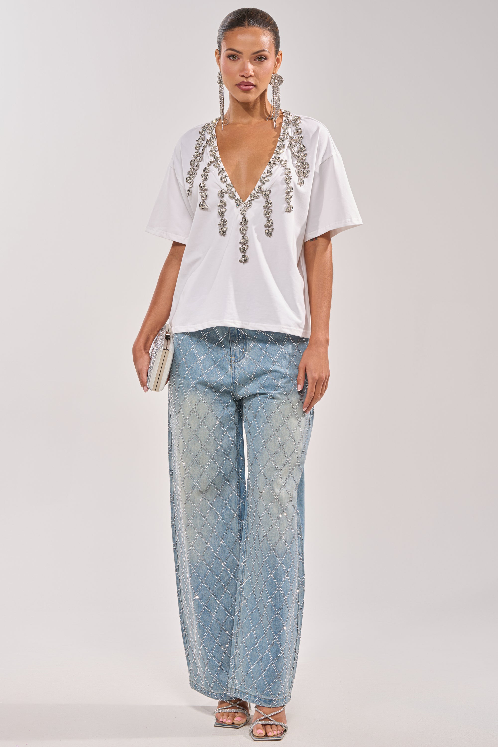A woman wears the TOO DEEP RHINESTONE TEE, wide-leg light blue diamond-pattern jeans, silver strappy heels, large earrings, and holds a small white purse. 8