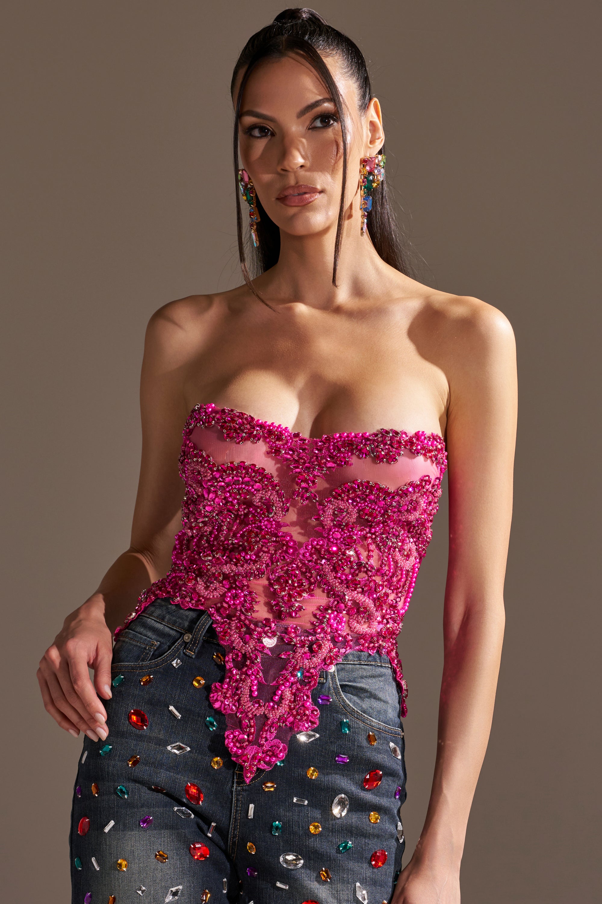 A woman poses confidently against a neutral background, wearing the OH HOT DAMN RHINESTONE CORSET and high-waisted, gem-embellished jeans.