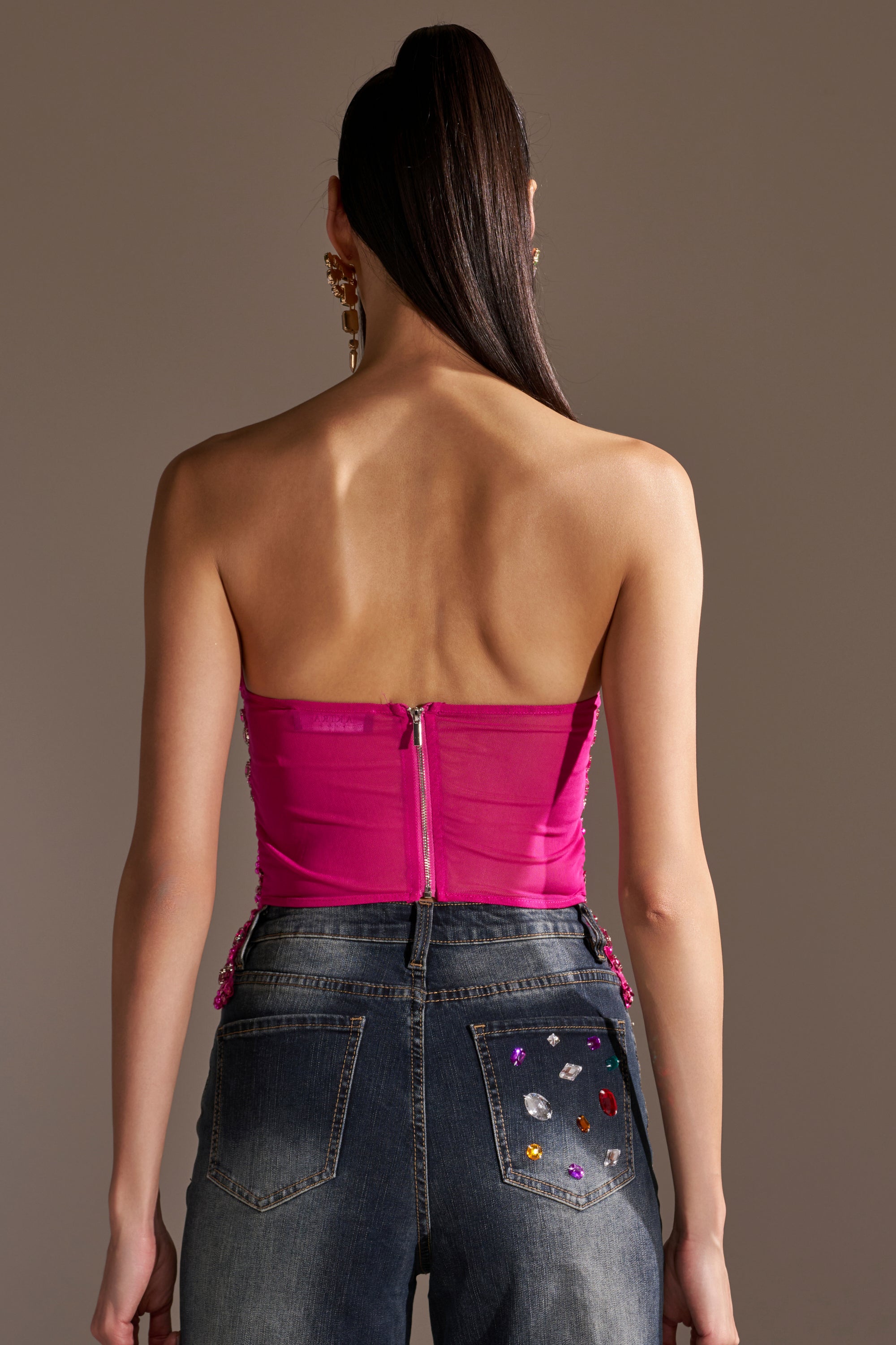 A woman with long dark hair in a high ponytail is shown from the back, wearing the OH HOT DAMN RHINESTONE CORSET in magenta and high-waisted blue jeans with colorful gems on the right pocket.