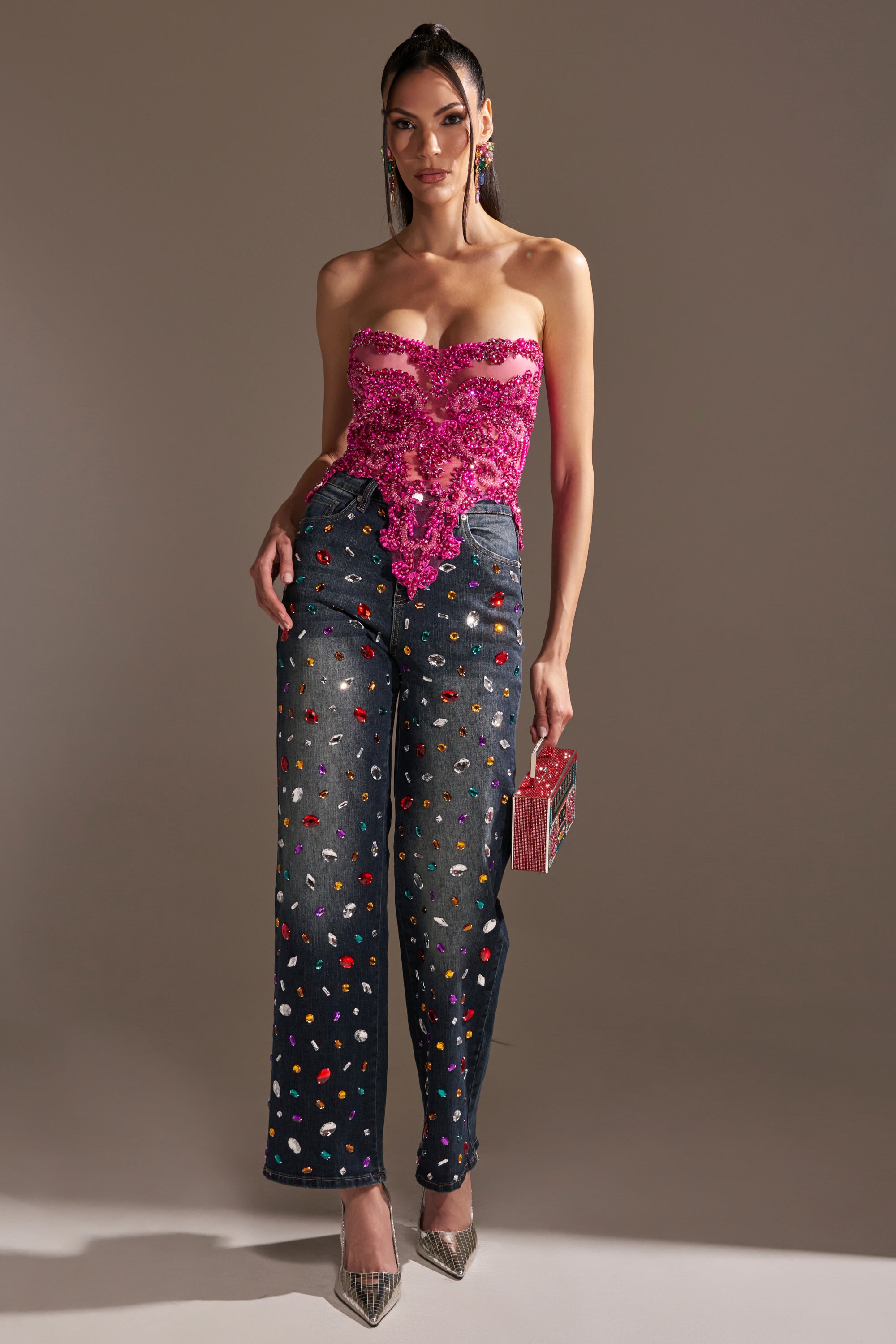 A woman stands against a plain background, wearing the OH HOT DAMN RHINESTONE CORSET and high-waisted, wide-leg embellished jeans. She holds a box-shaped handbag and wears silver heels.