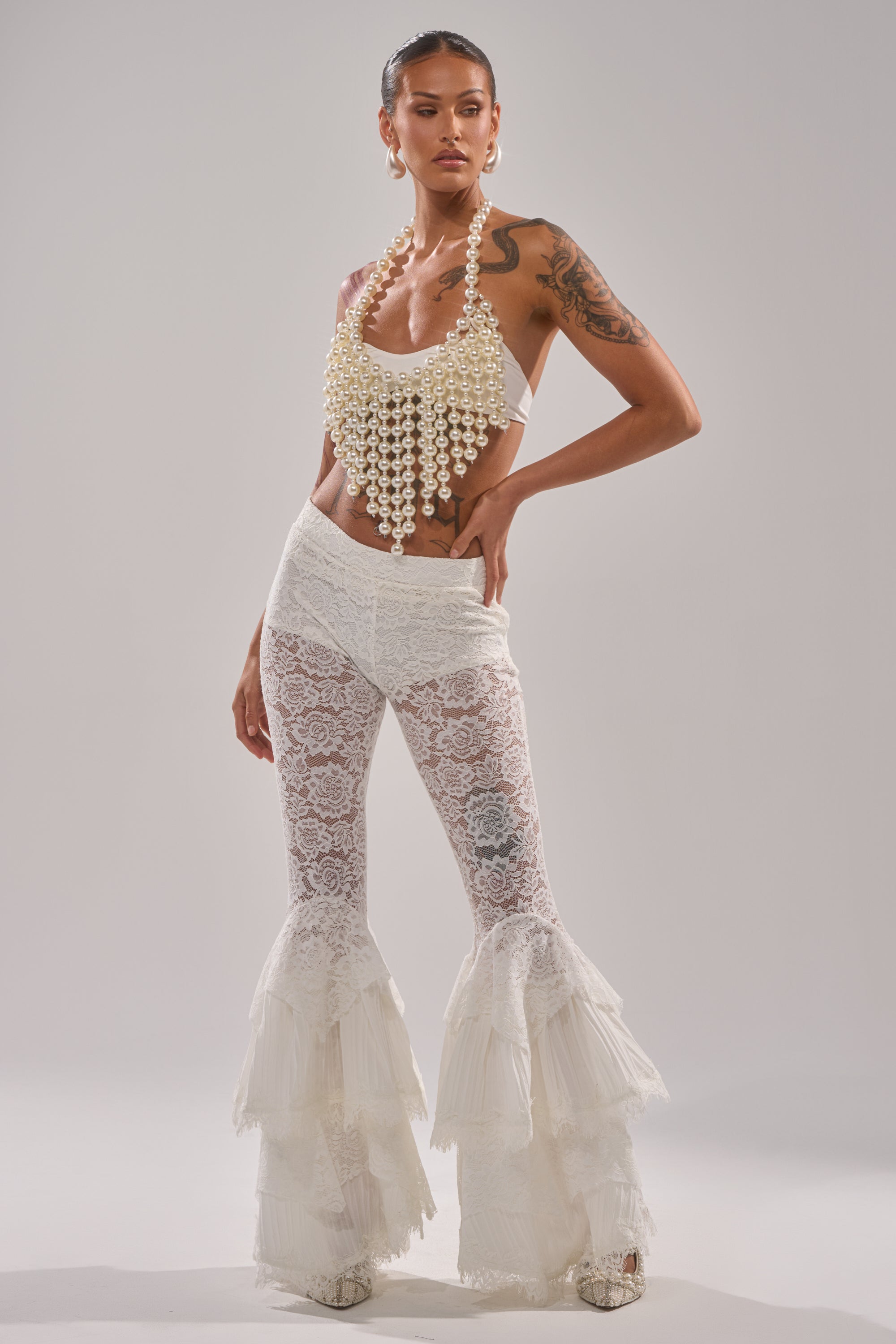 A woman confidently models the QUEEN SH*T BRALETTE, styled with white lace flared pants featuring layered ruffles and hoop earrings. Against a plain background, she displays her tattoos and bold fashion sense.