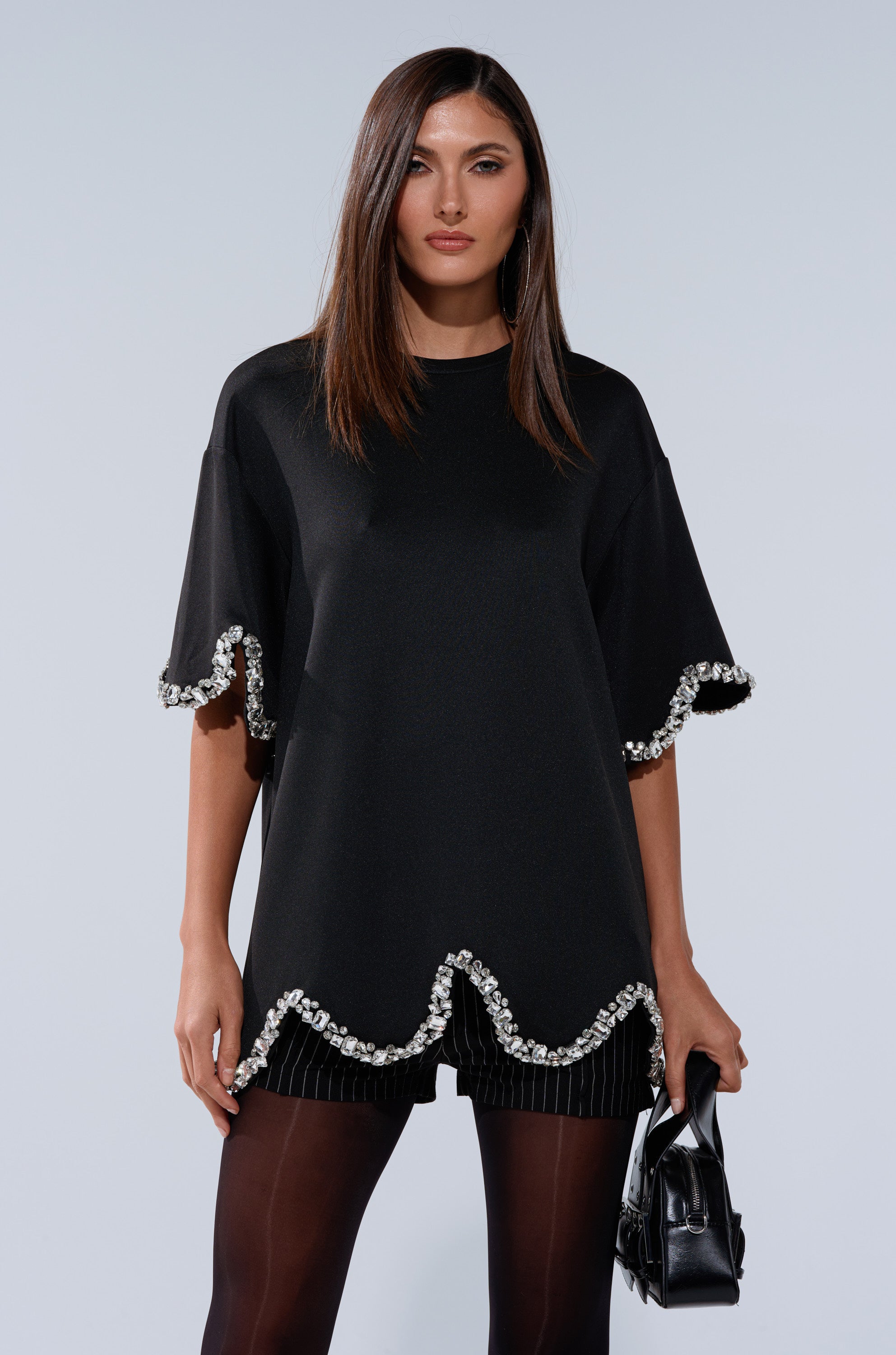 A woman wears the AT LEAST I'M ALIVE OVERSIZED TEE WITH RHINESTONES, paired with black shorts, tights, and a small black handbag, as she stands against a plain light background.