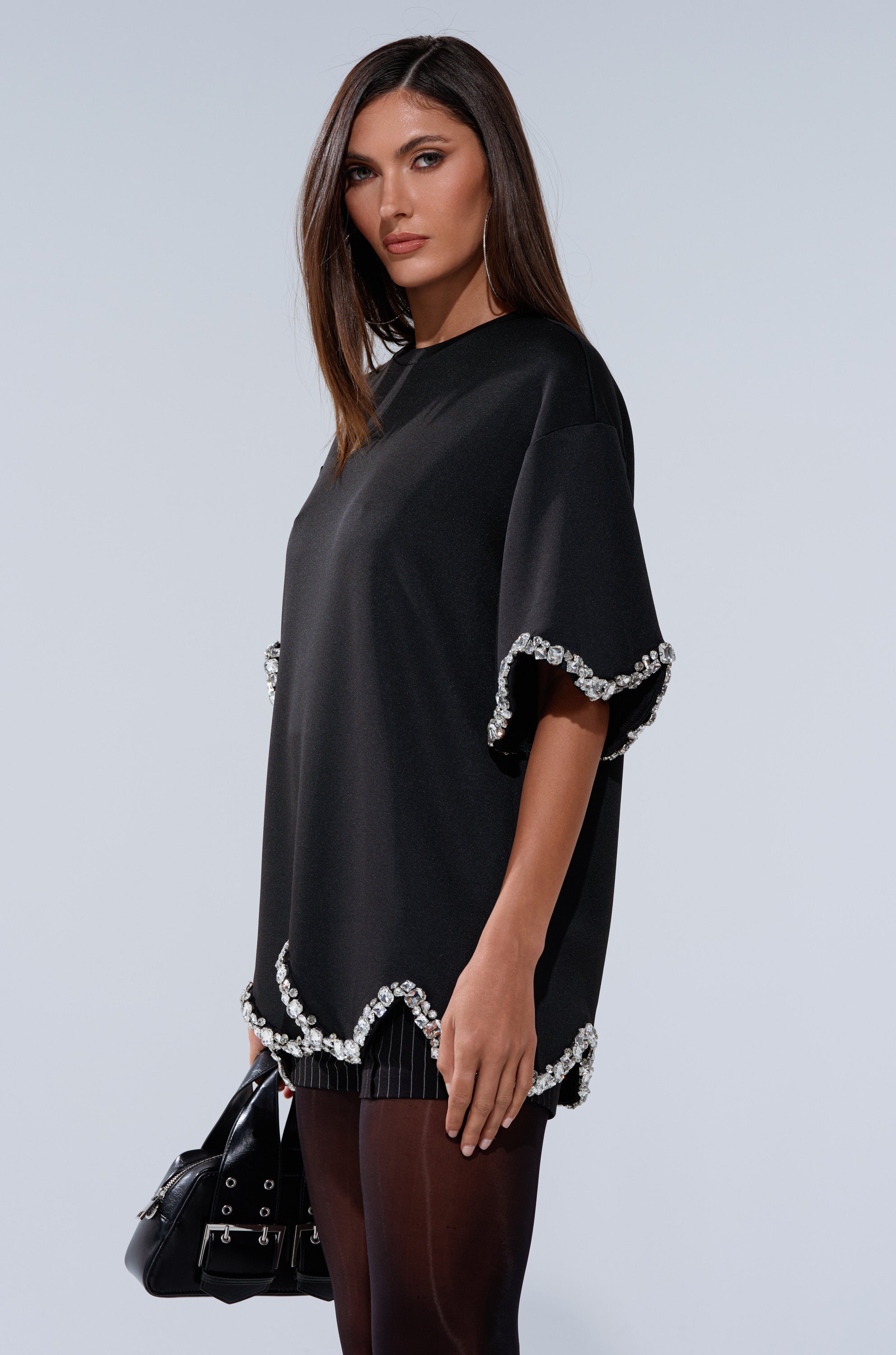 A woman with straight brown hair wears the "AT LEAST I'M ALIVE OVERSIZED TEE WITH RHINESTONES," a loose black dress with rhinestone-trimmed sleeves and hem, paired with sheer black tights and a black handbag, against a light gray background.