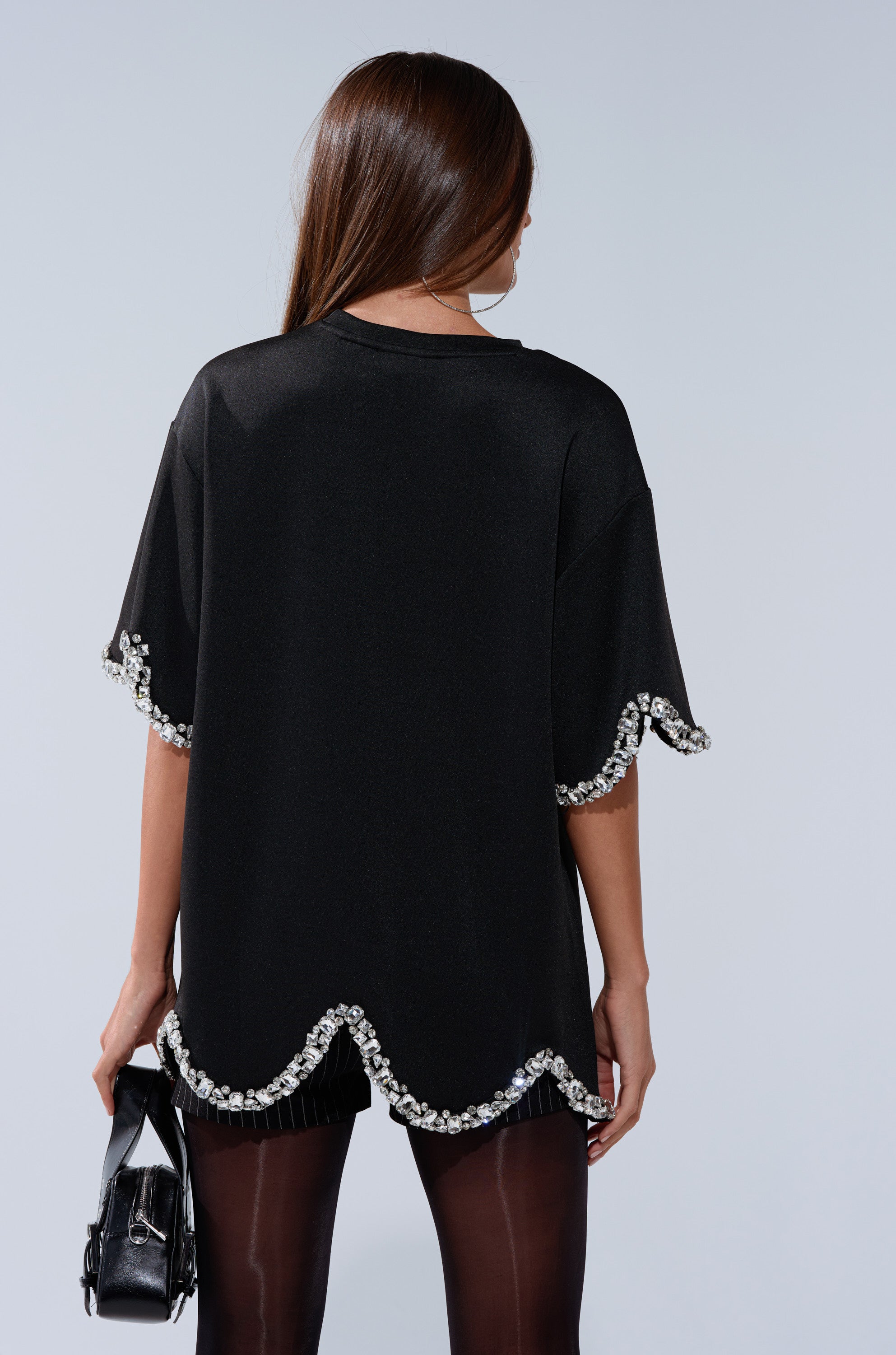 A woman with long brown hair, seen from behind, wears the AT LEAST I'M ALIVE OVERSIZED TEE WITH RHINESTONES— a black, streetwear-inspired top with scalloped hem and rhinestone-trimmed sleeves. She holds a small black handbag and wears black tights.