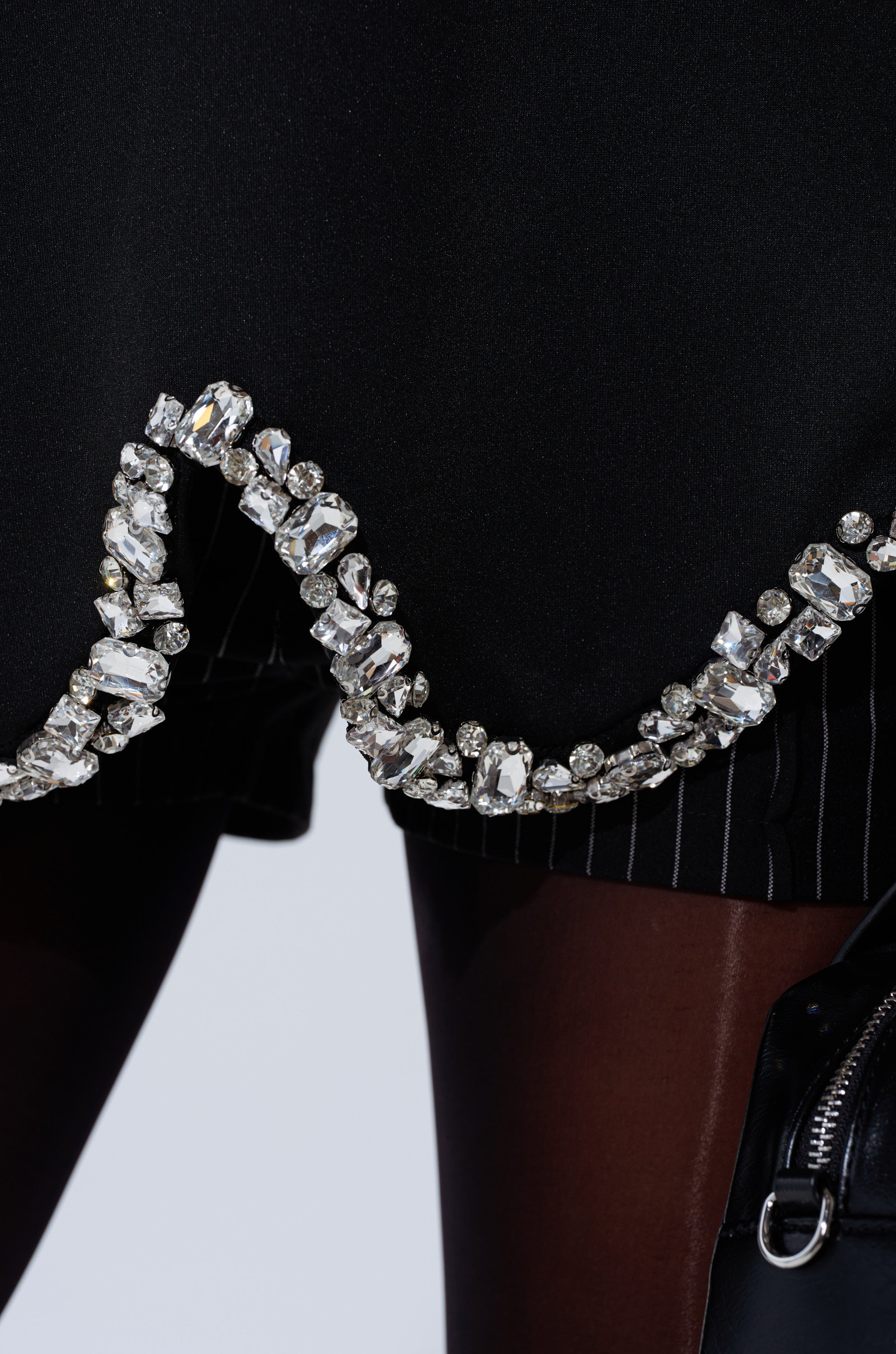 Close-up of the AT LEAST I'M ALIVE OVERSIZED TEE WITH RHINESTONES, a black tee featuring a wavy trim adorned with large sparkling rhinestones. Background shows sheer black tights and part of a streetwear-inspired black leather accessory.