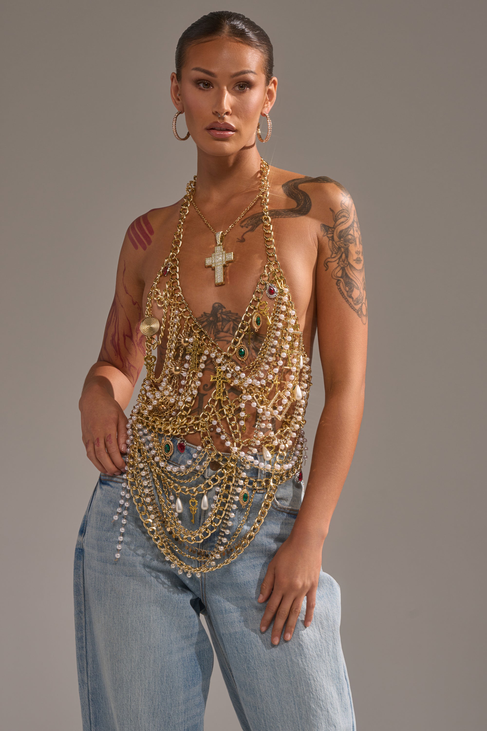 A person models the DRIP DRIP BRALETTE—gold chain style layered with pearl necklaces—as a standout festival top, paired with light blue jeans, hoop earrings, and styled hair against a gray background. Tattoos are visible. 0