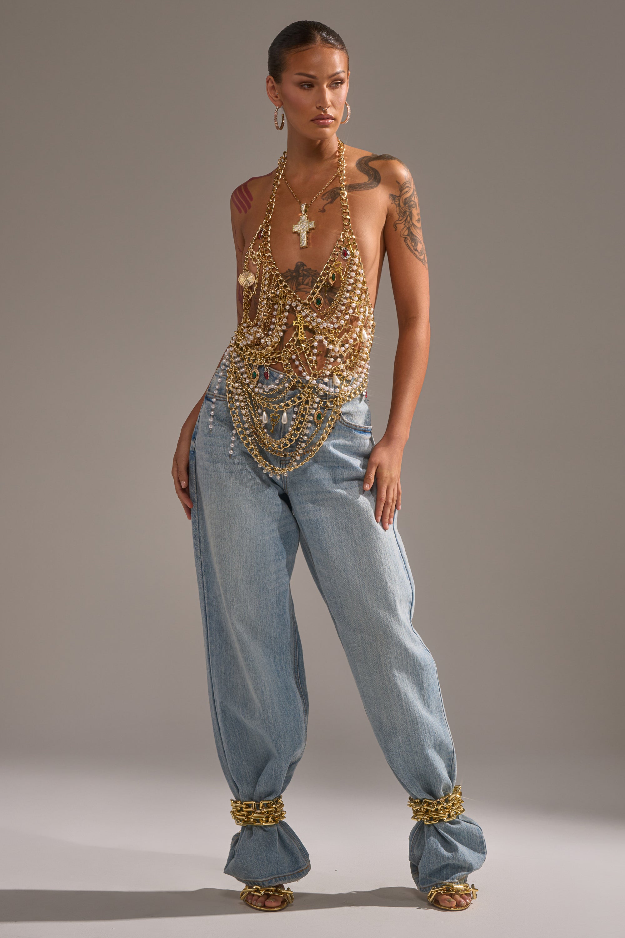 A woman poses confidently in light blue baggy jeans and the DRIP DRIP BRALETTE, layered with gold jewelry on her neck, wrists, and ankles. Tattooed arms add to her bold look against a neutral gray background. 5