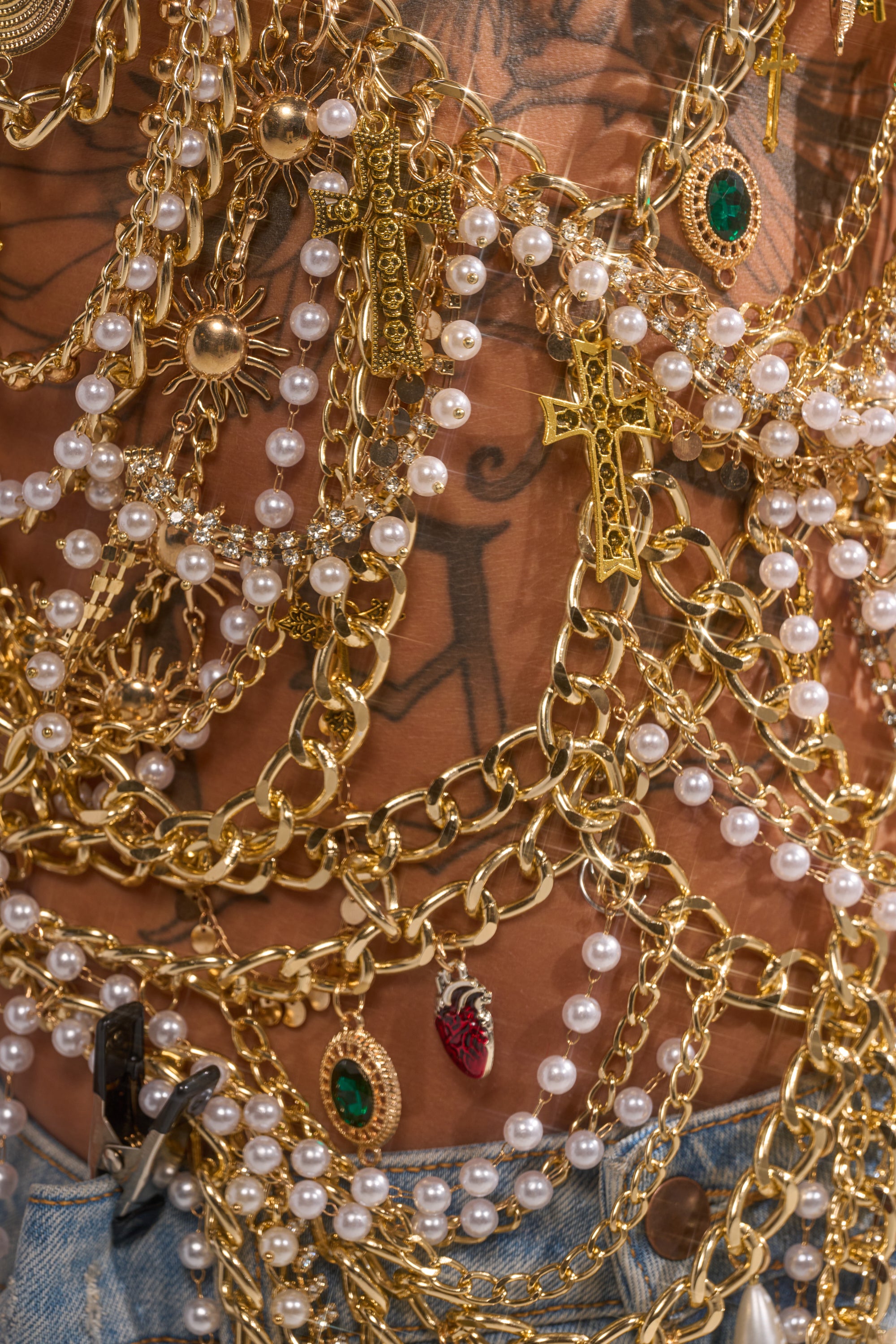 Close-up of the DRIP DRIP BRALETTE: layered gold chains and pearl necklaces with unique pendants draped over a tattooed torso, styled on blue denim for a bold, statement festival look. 8