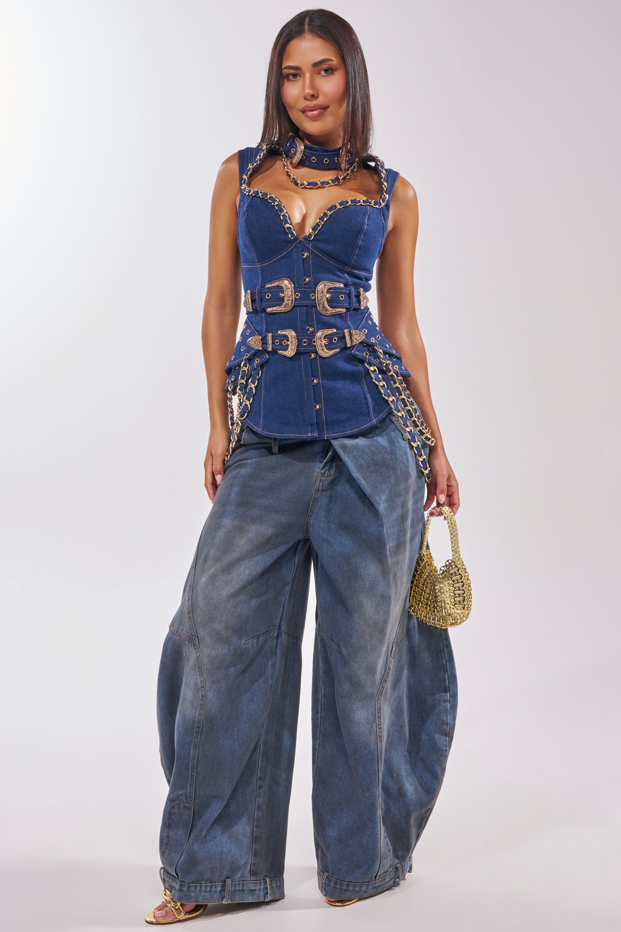 A woman stands confidently in the WHAT IN TARNATION DENIM CORSET, featuring sleeveless denim with chain details and paired with wide-leg jeans—ideal for festivals. She adds a gold handbag, heeled sandals, and poses against a neutral background. 5