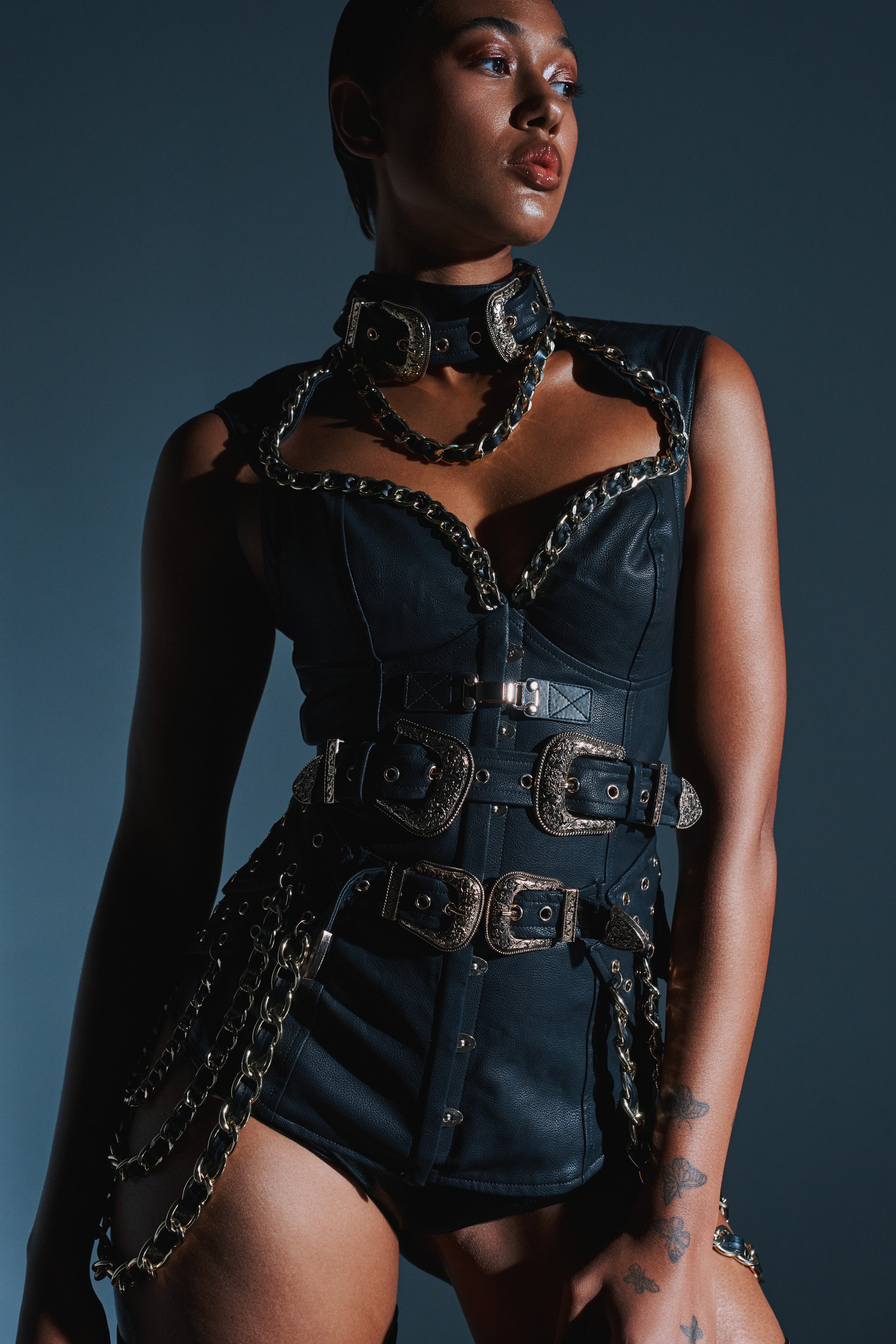 A person models the LETS GO TO THE RODEO BLOUSE—black vegan leather with chain and buckle accents—against a dark backdrop, sporting short hair, bold makeup, and tattoos for an edgy, gothic rodeo vibe. 0