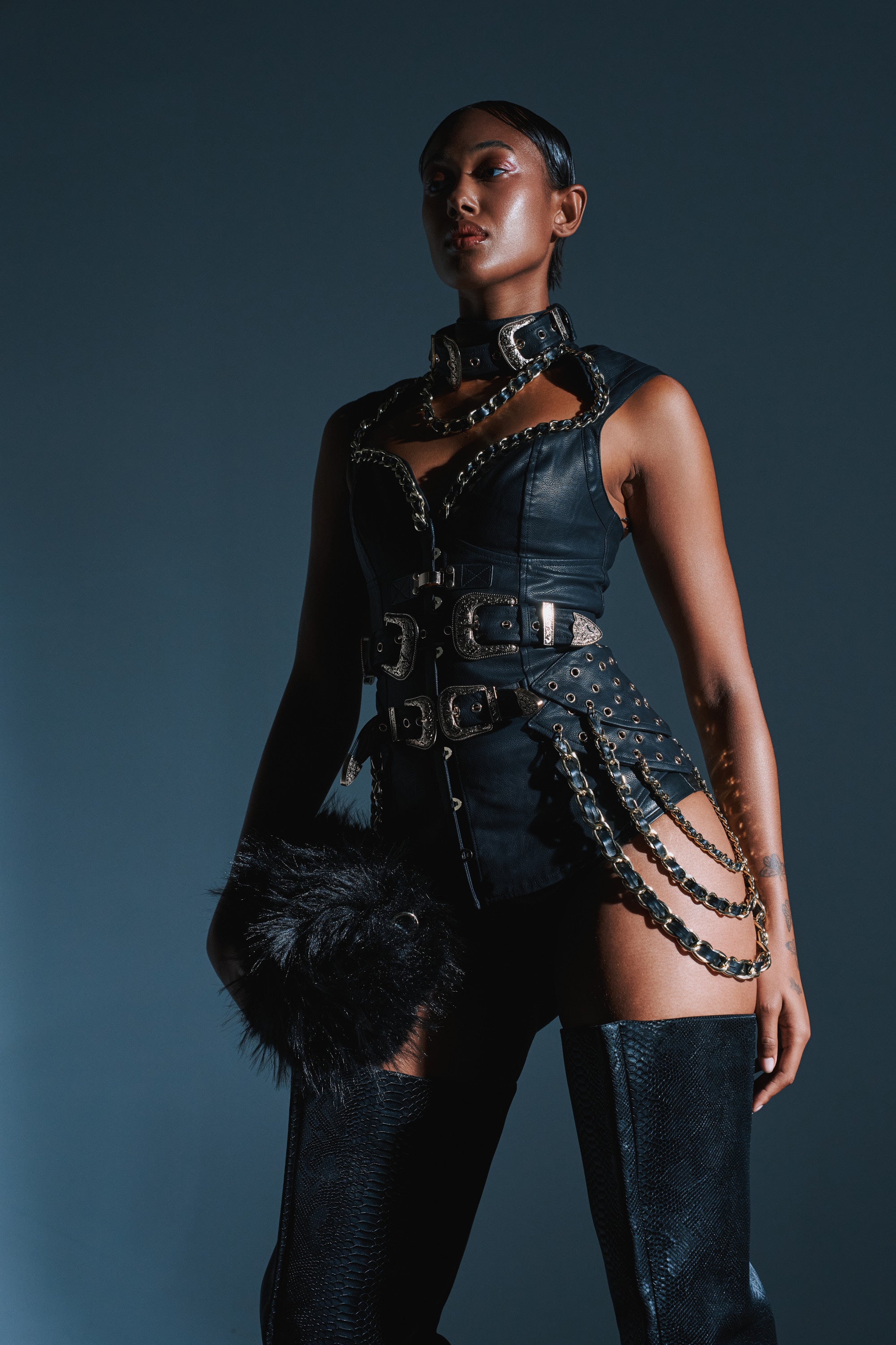 A woman poses confidently in the LETS GO TO THE RODEO BLOUSE, a black vegan leather top featuring gold chains and multiple buckles, paired with high black boots and holding a black fur accessory against a dark backdrop. 1