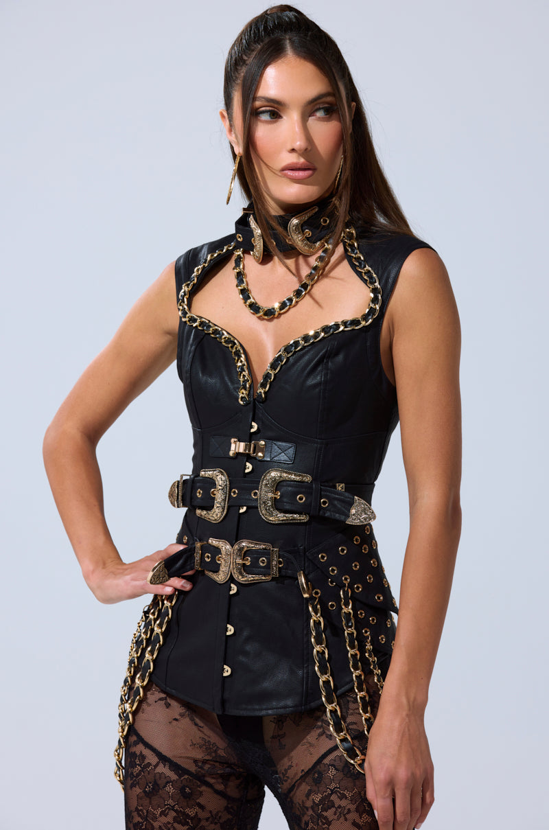 She wears the LETS GO TO THE RODEO BLOUSE, a fitted black corset-style blouse dress with gold chain, buckle, and grommet details, layered over black lace tights. Her long brown hair is styled back and she sports bold makeup. 5