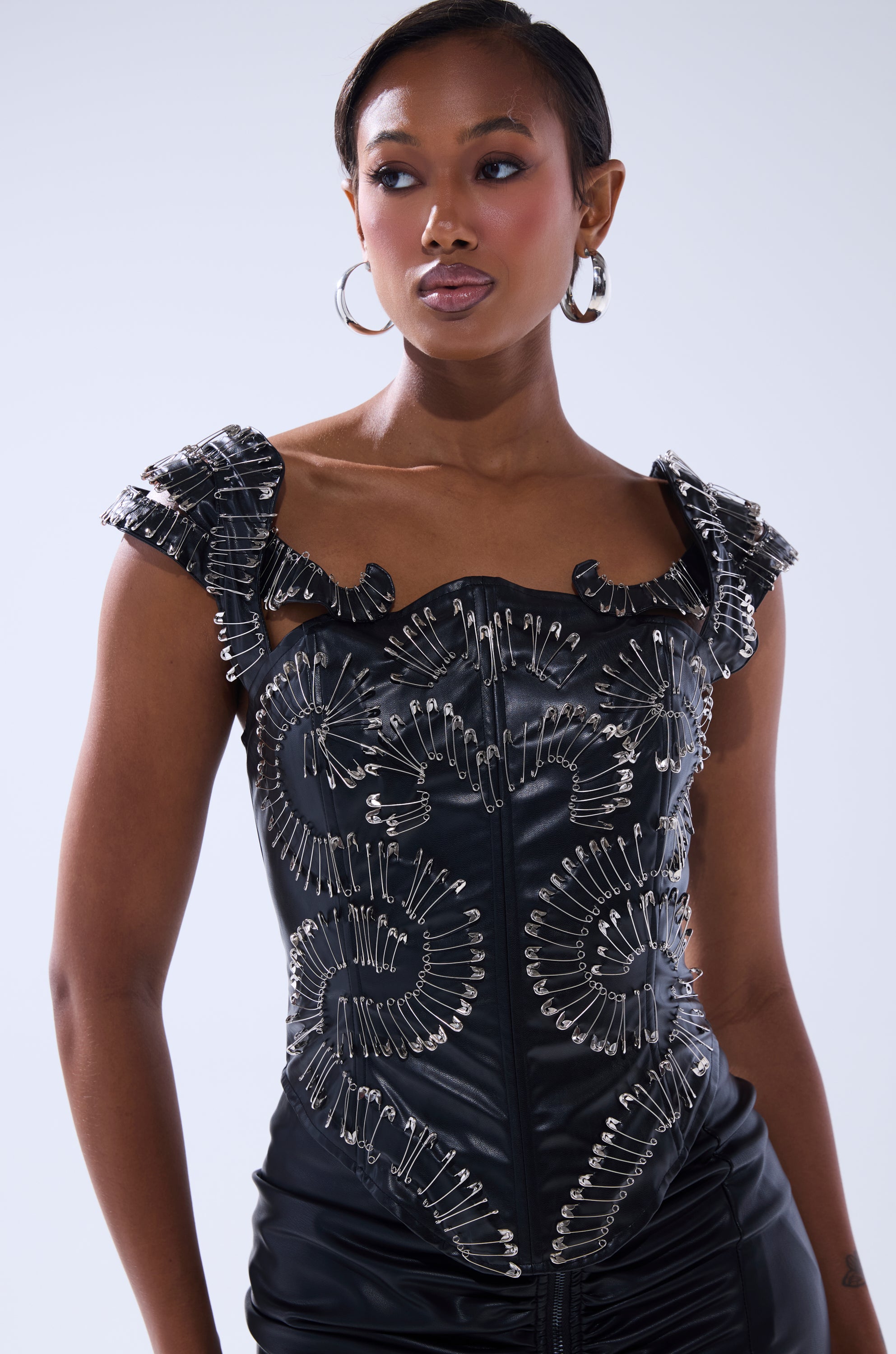 A woman models the PIN ME DOWN FAUX LEATHER CORSET, a black corset embellished with decorative silver pins, paired with matching black bottoms against a plain light background.