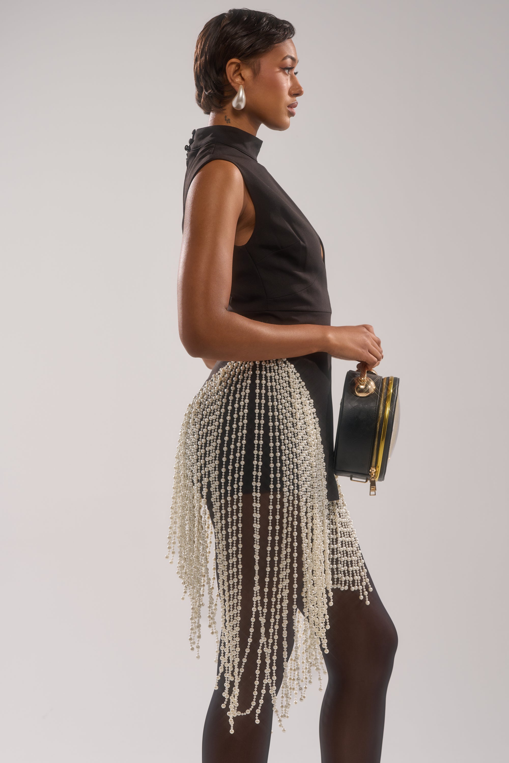 A woman in profile wears the TOGETHER FOREVER BLOUSE WITH PEARLS, black shorts, sheer tights, and a beaded fringe skirt. She accessorizes with a black clutch and large round earrings against a neutral background.