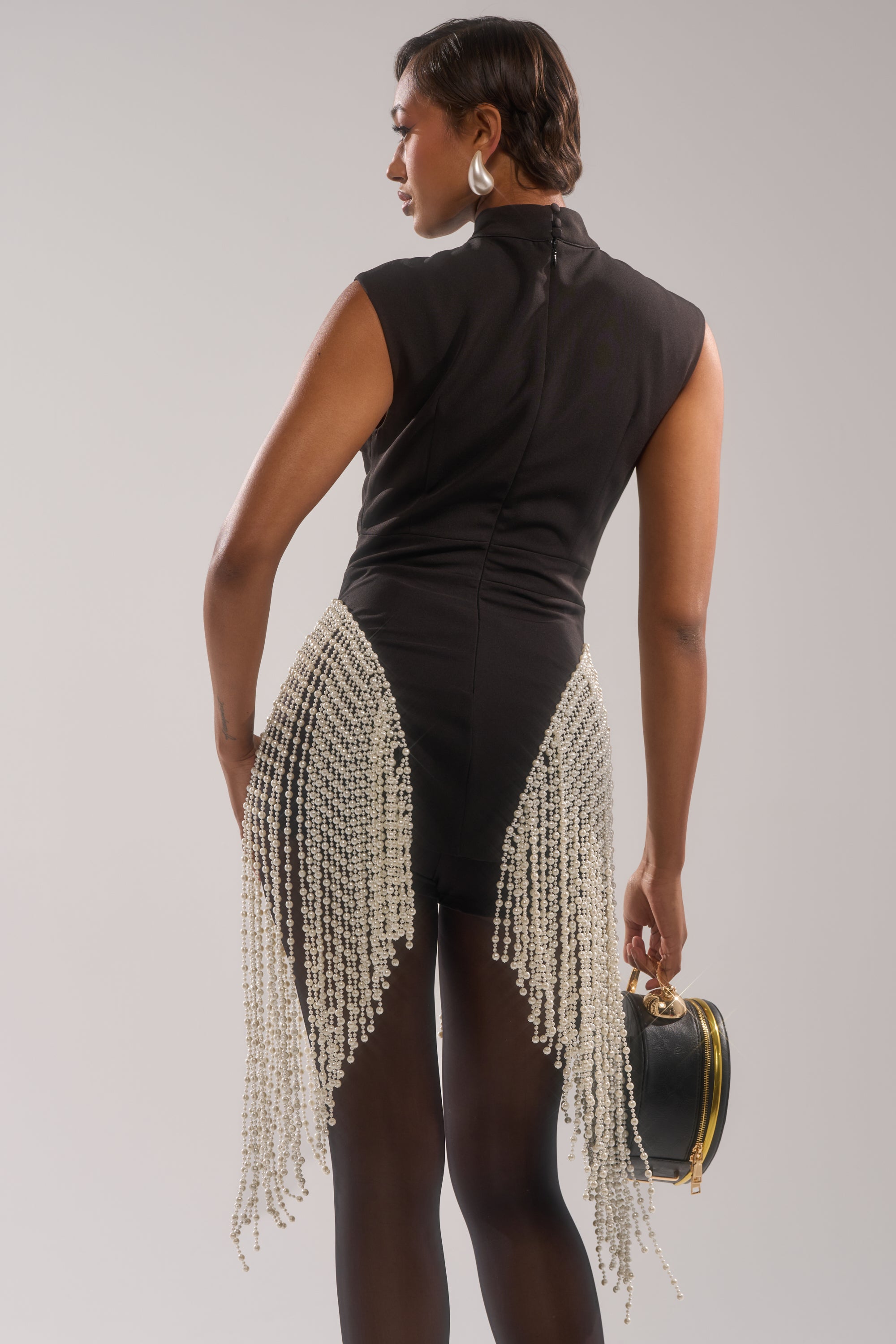 Wearing the TOGETHER FOREVER BLOUSE WITH PEARLS, a person pairs it with black tights, silver earrings, and a black handbag while posing slightly turned against a neutral background.
