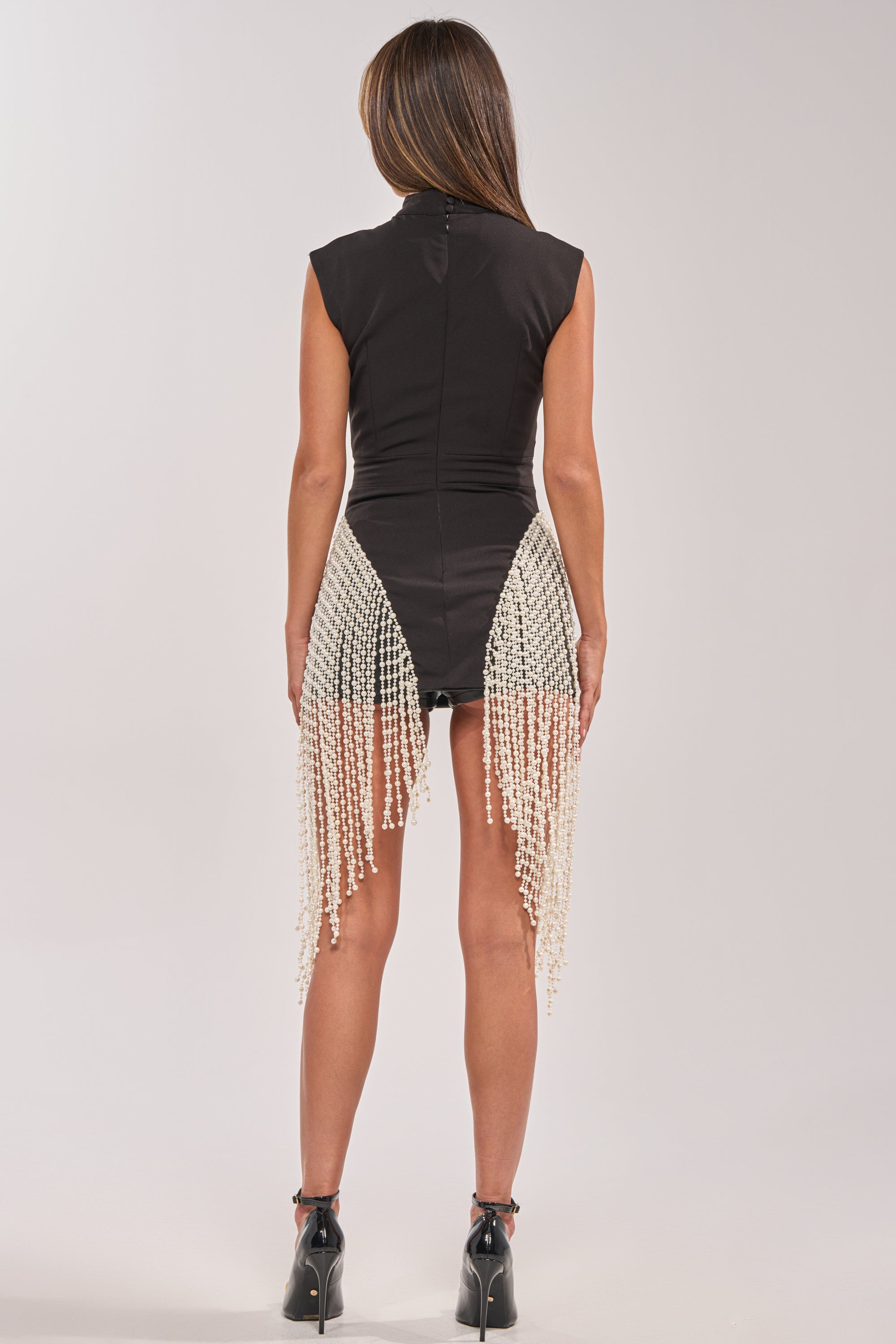 A woman faces away, wearing the TOGETHER FOREVER BLOUSE WITH PEARLS—fitted, sleeveless, black with pearl fringe details—paired with black heeled ankle boots against a plain light gray background. 3