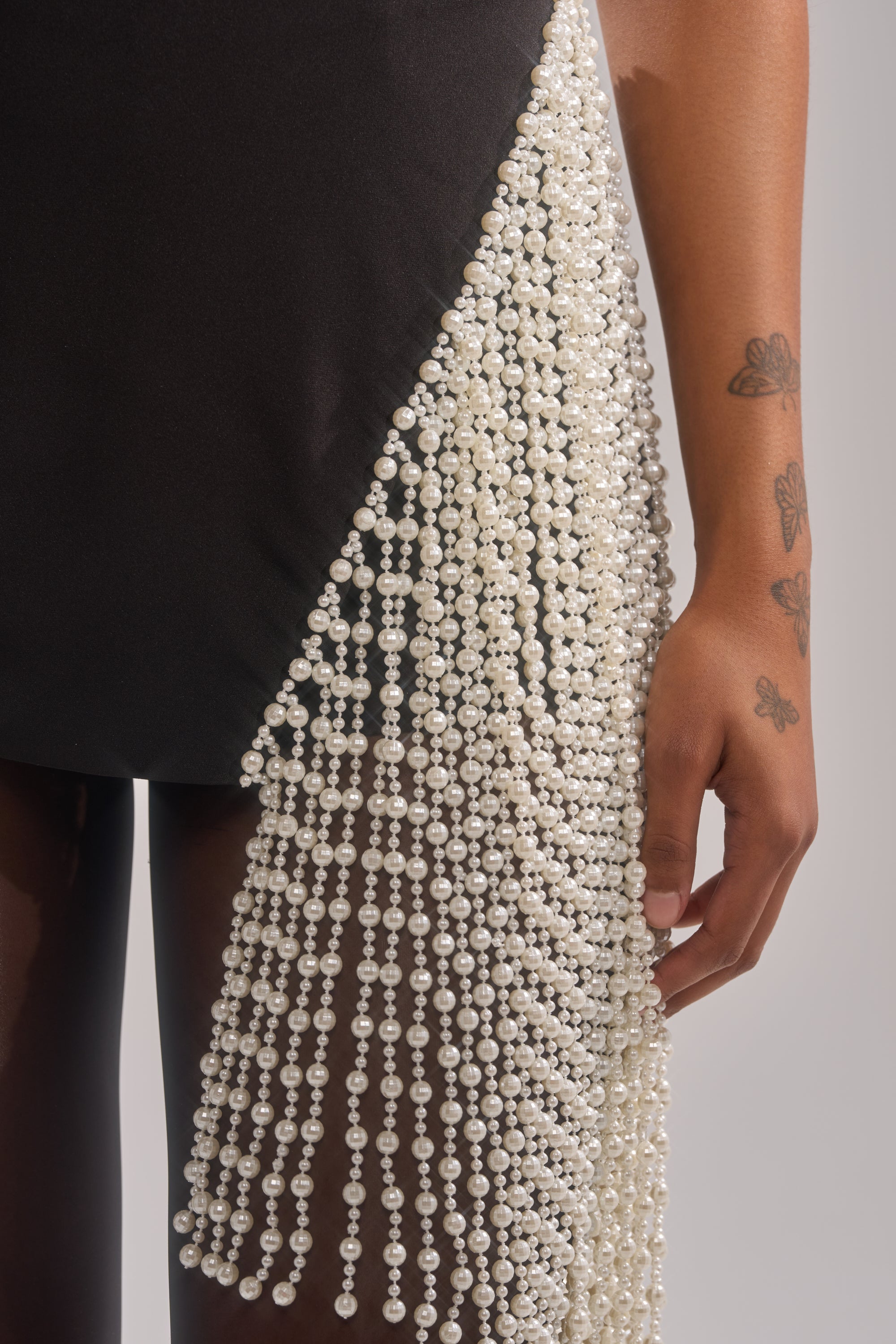 Wearing the TOGETHER FOREVER BLOUSE WITH PEARLS, a black sleeveless fitted top with cascading pearl bead fringe, a person with butterfly tattoos on their arm is shown in a cropped image featuring their lower torso and arm.