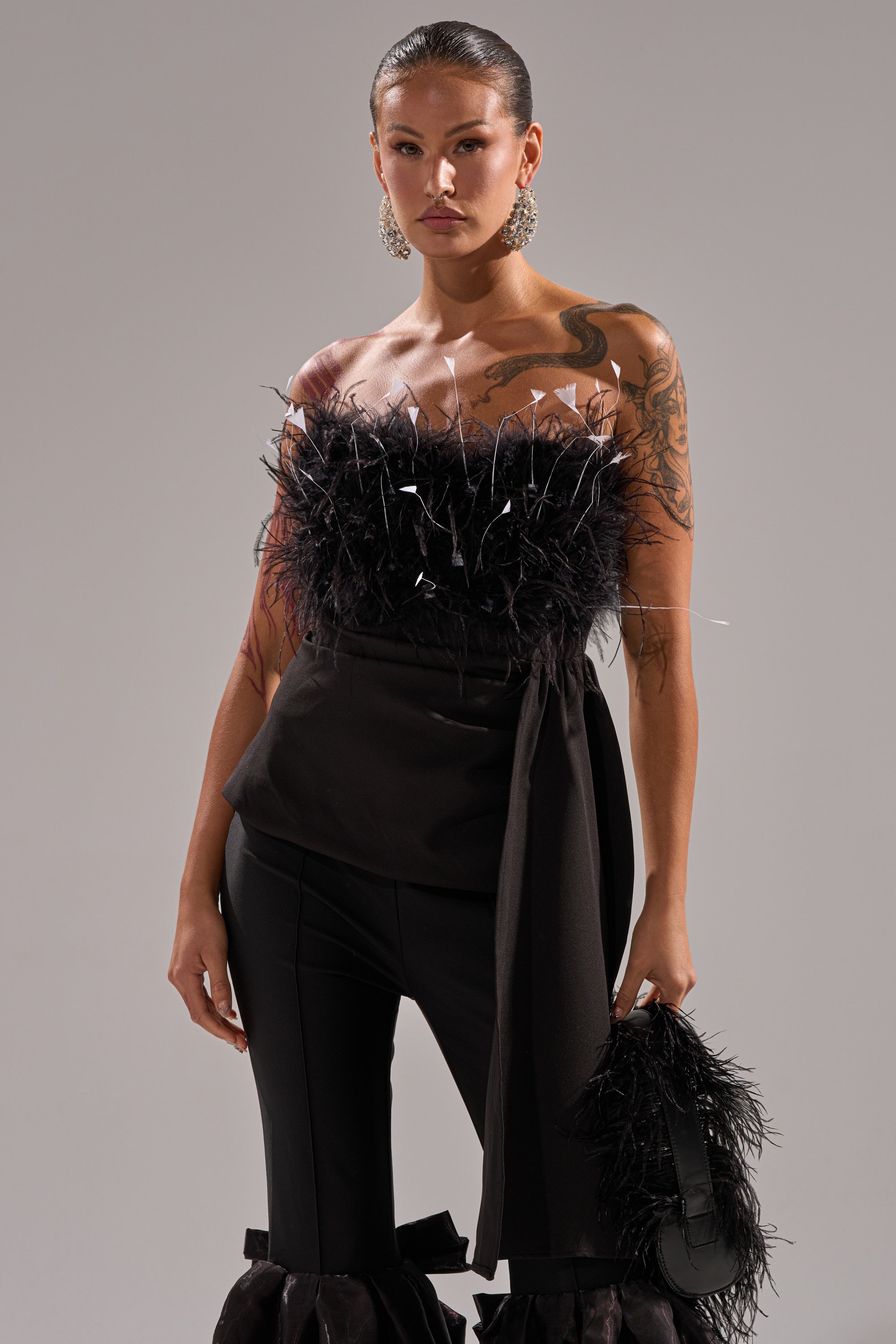 A woman with slicked-back hair wears the CLARA FEATHER CORSET, styled with matching black pants and statement earrings. She carries a black feathered clutch and displays tattoos on her arm.
