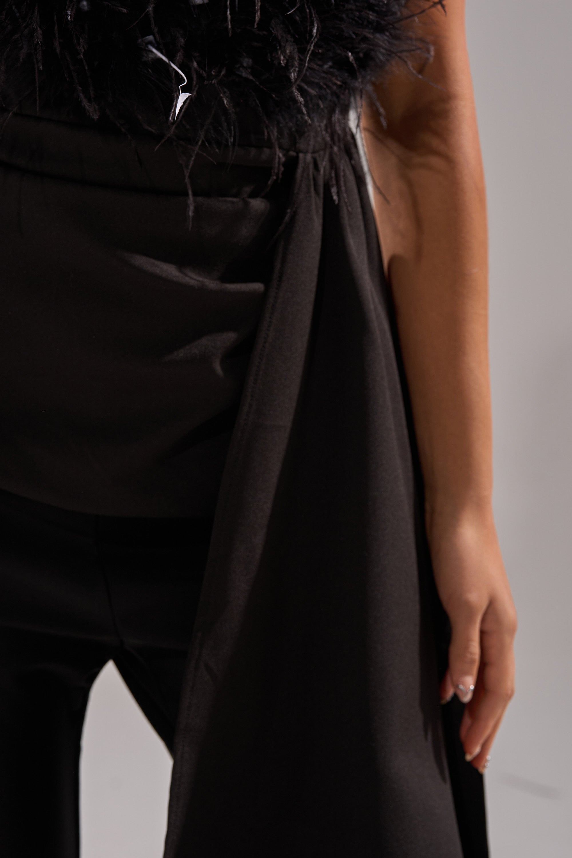 A close-up of a person wearing black pants and the CLARA FEATHER CORSET, their right arm relaxed by their side against a plain light background—an elegant look ideal for a night out.