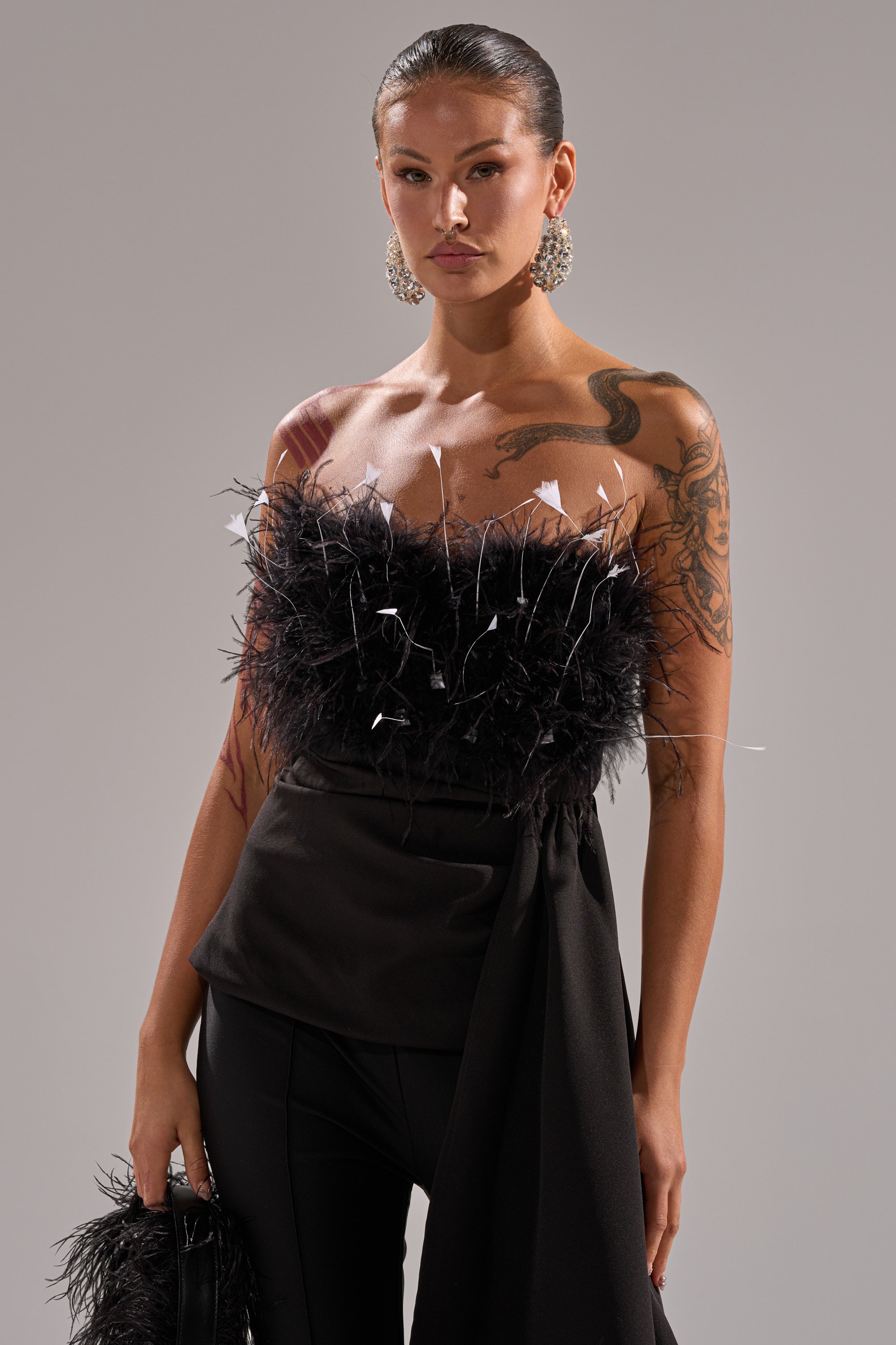 A woman with tattoos wears the CLARA FEATHER CORSET—strapless, black, with a feather trim—paired with black pants and bold earrings. She holds a matching bag and poses confidently against a gray background.