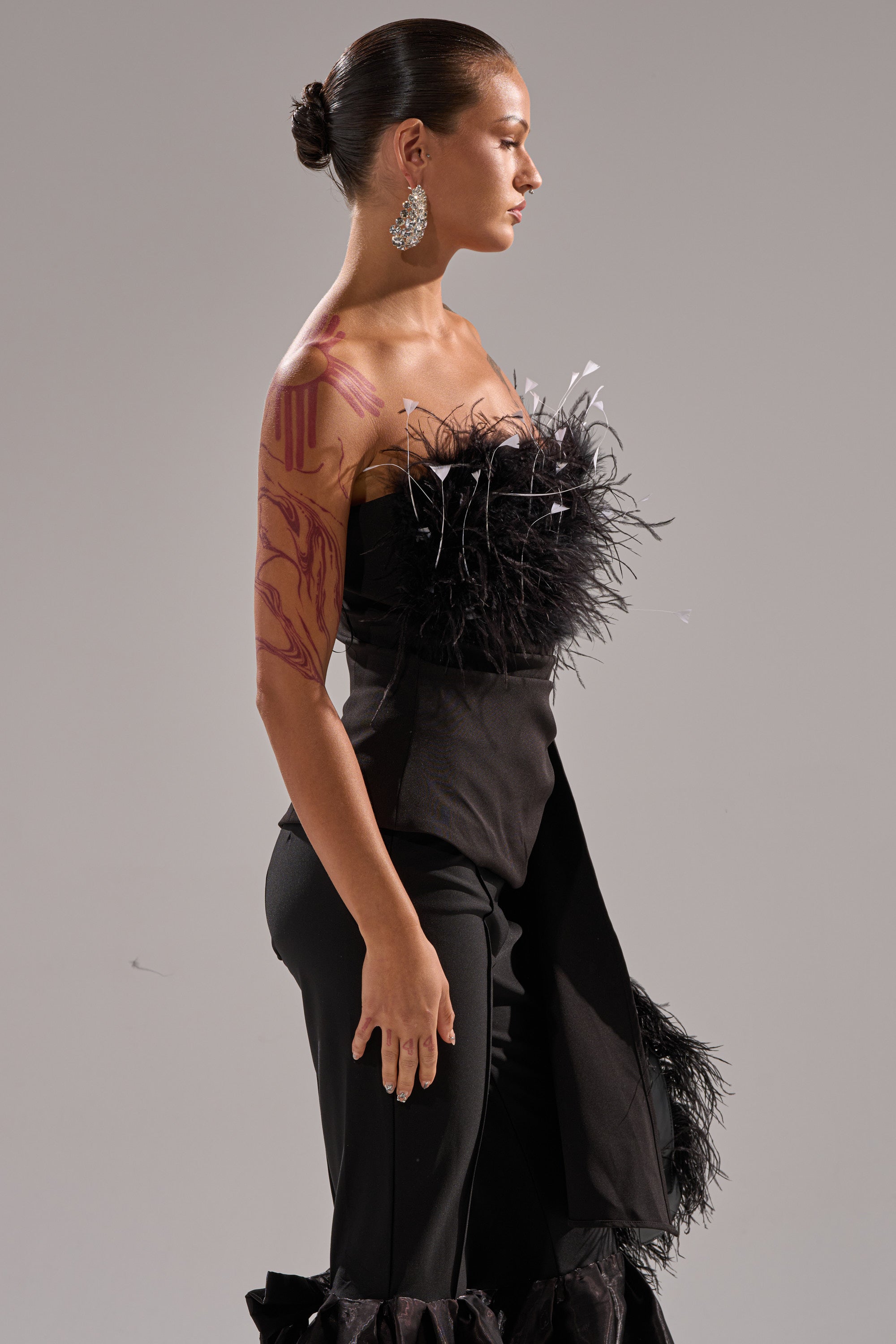 A woman stands in profile wearing the CLARA FEATHER CORSET with a strapless neckline and ruffled pants. She has slicked-back hair in a bun, large dangling earrings, and red designs painted on her right arm and shoulder.