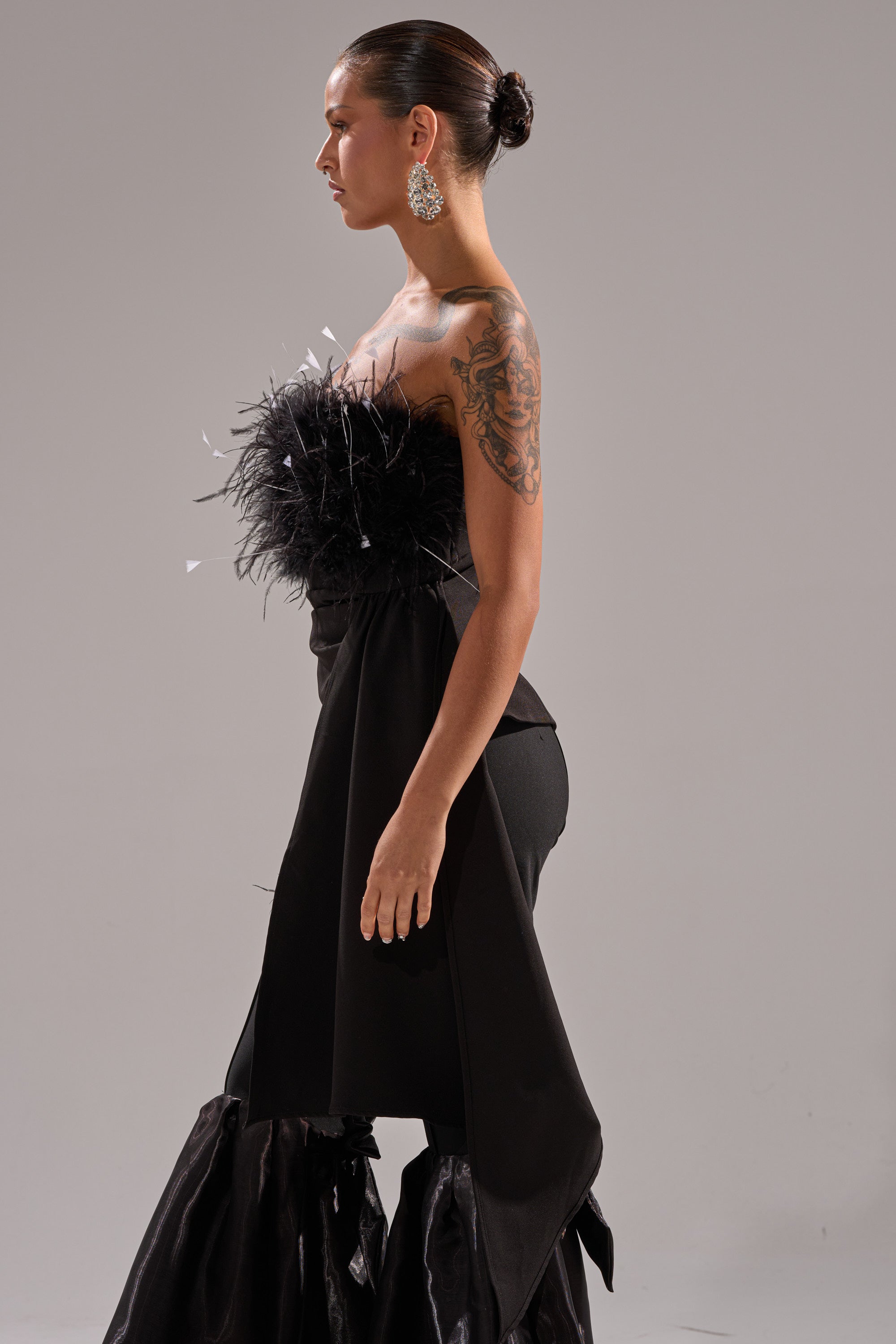 A woman with a sleek bun and large earrings stands in profile, wearing the CLARA FEATHER CORSET with a strapless neckline, feather detailing, and ruffles. She displays an arm tattoo and maintains a neutral expression.