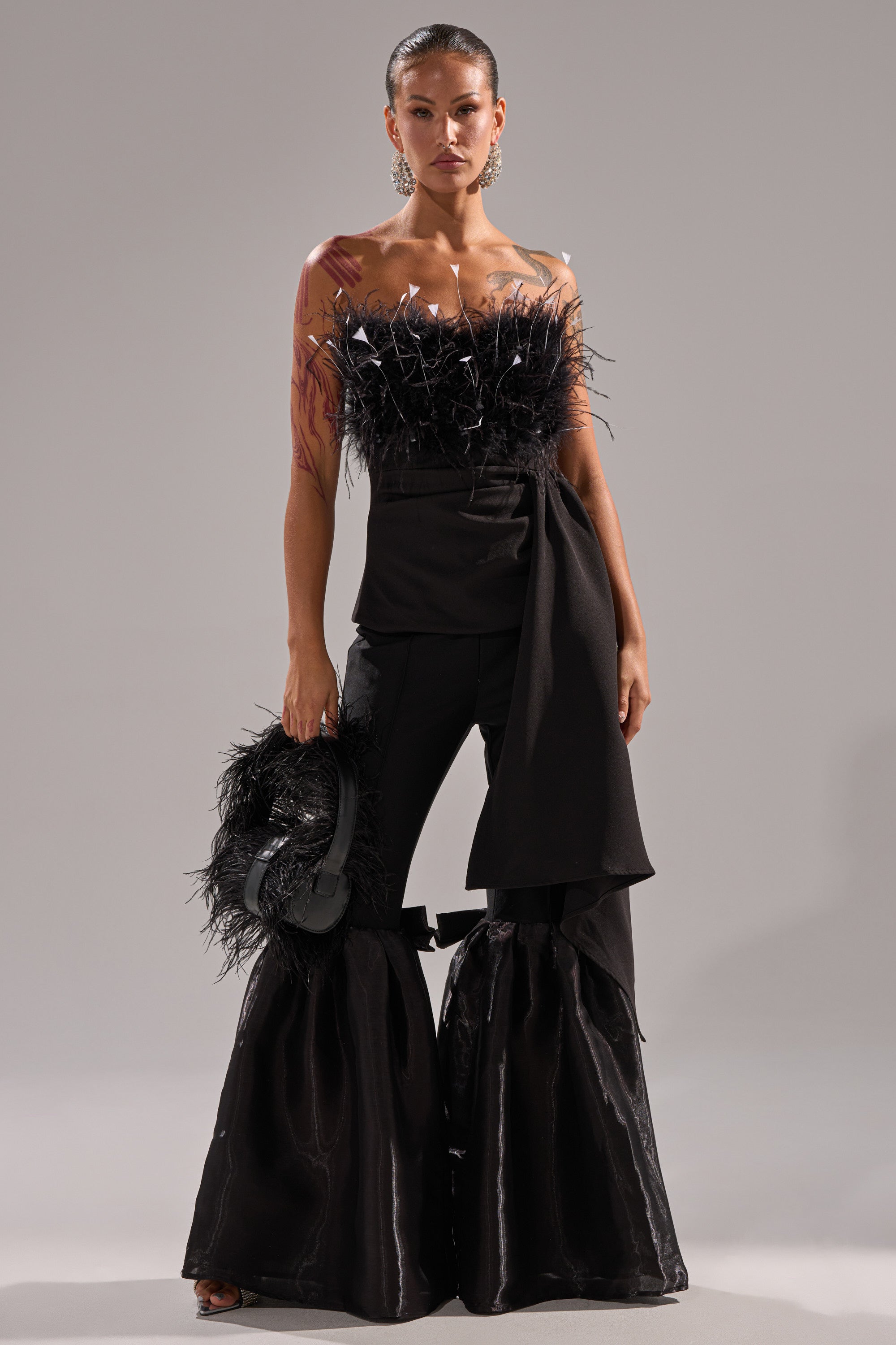 A woman stands against a plain background wearing the CLARA FEATHER CORSET, a strapless night-out top with feather details. She pairs it with black flared pants featuring shiny knee panels, holds a feathered accessory, and wears statement earrings.