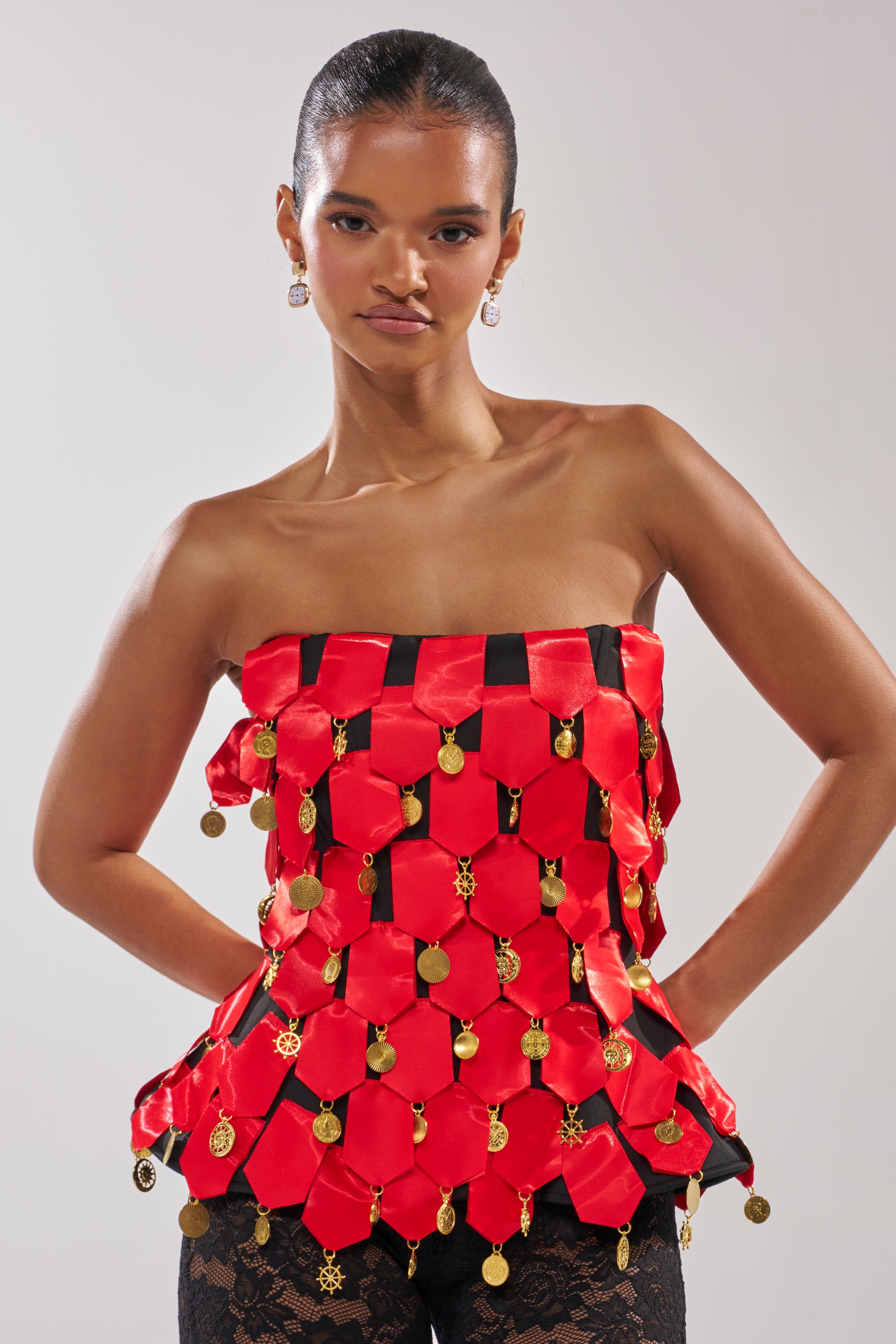 A woman stands confidently with her hands on her hips against a plain background, wearing the NOBODY KNOWS CORSET—a strapless red satin top adorned with gold coin-like charms—paired with black lace pants.