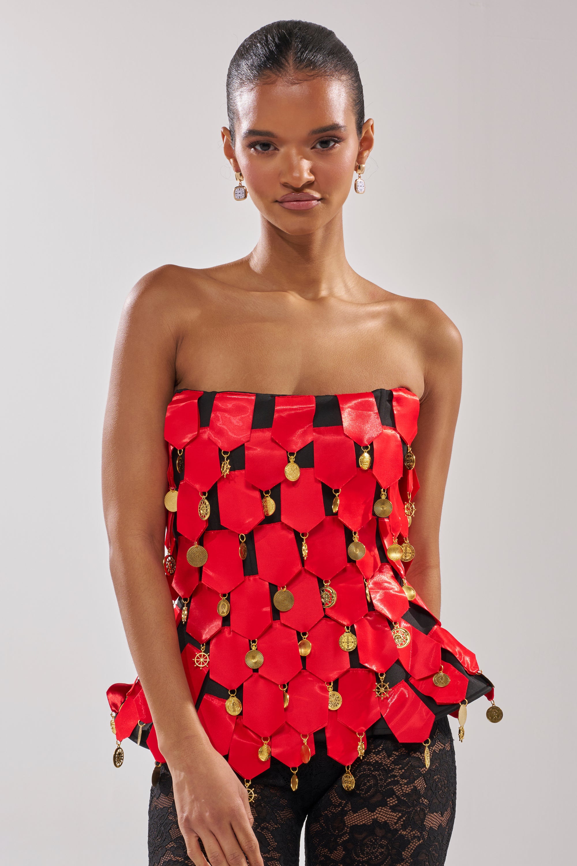 A woman models the NOBODY KNOWS CORSET, a strapless red satin piece with gold coin accents, paired with black lace pants against a plain light background.