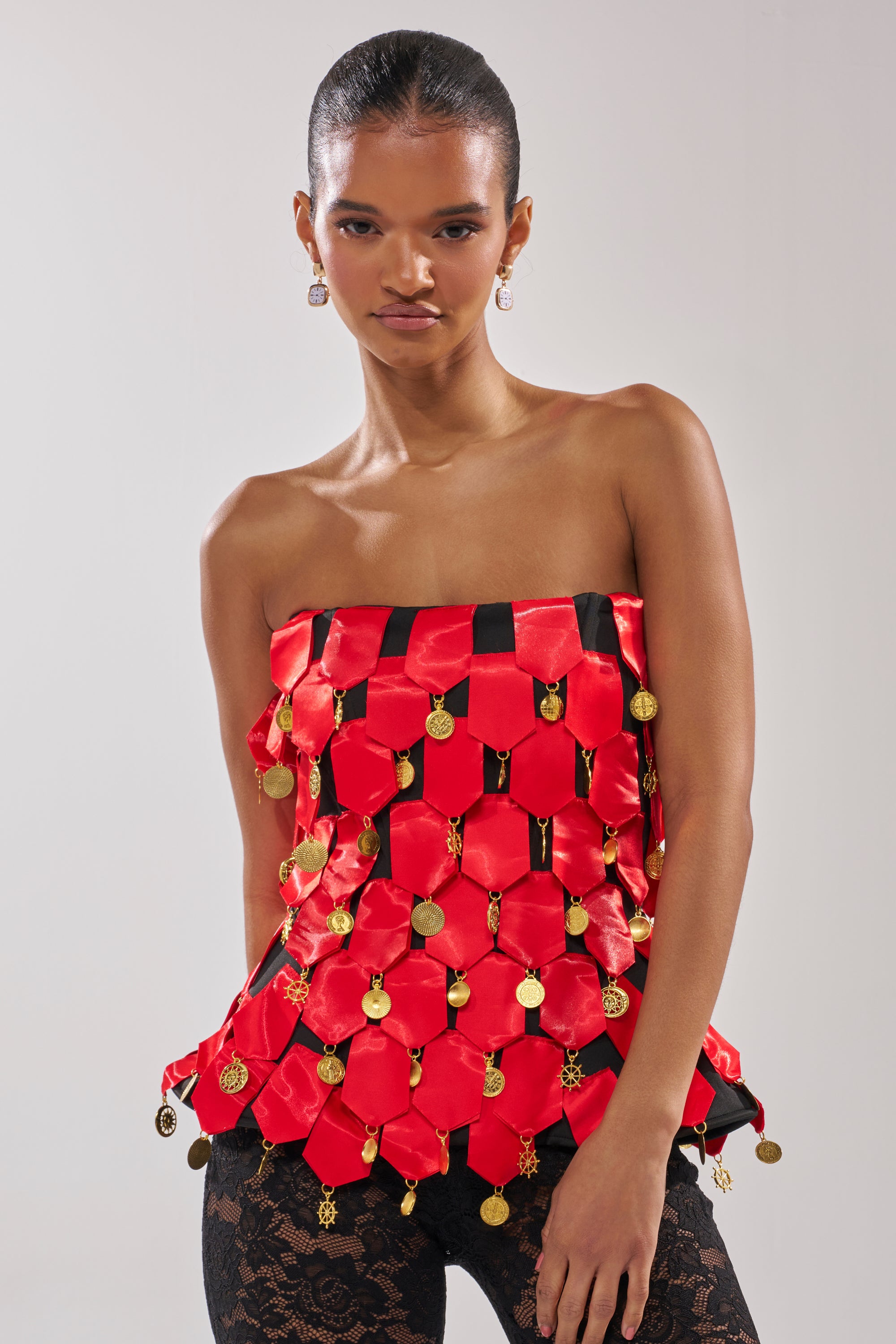 A woman wears the NOBODY KNOWS CORSET, a strapless red satin geometric top with gold coin-like charms, paired with black lace pants against a plain light background.