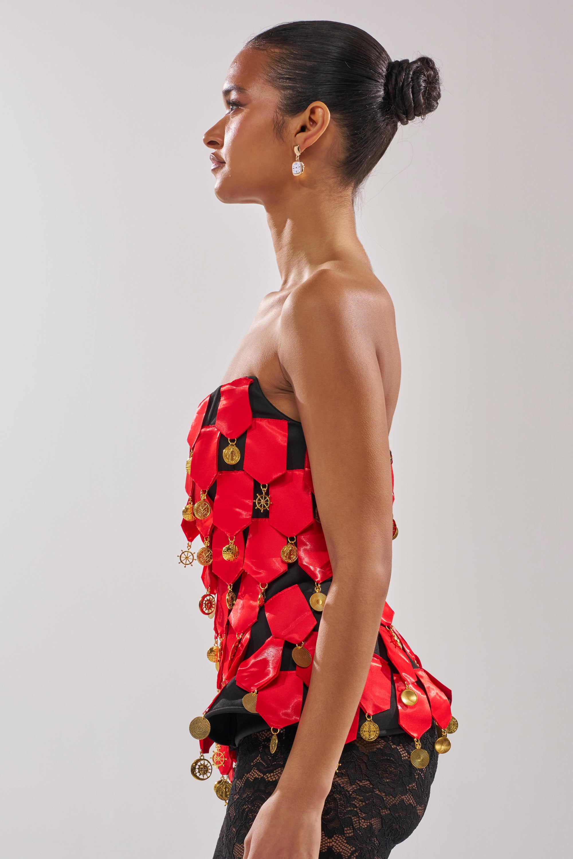 A woman with a neat bun and earrings models the NOBODY KNOWS CORSET, a red satin corset-style top adorned with gold coins, paired with patterned black lace pants, posing in profile against a plain light background.