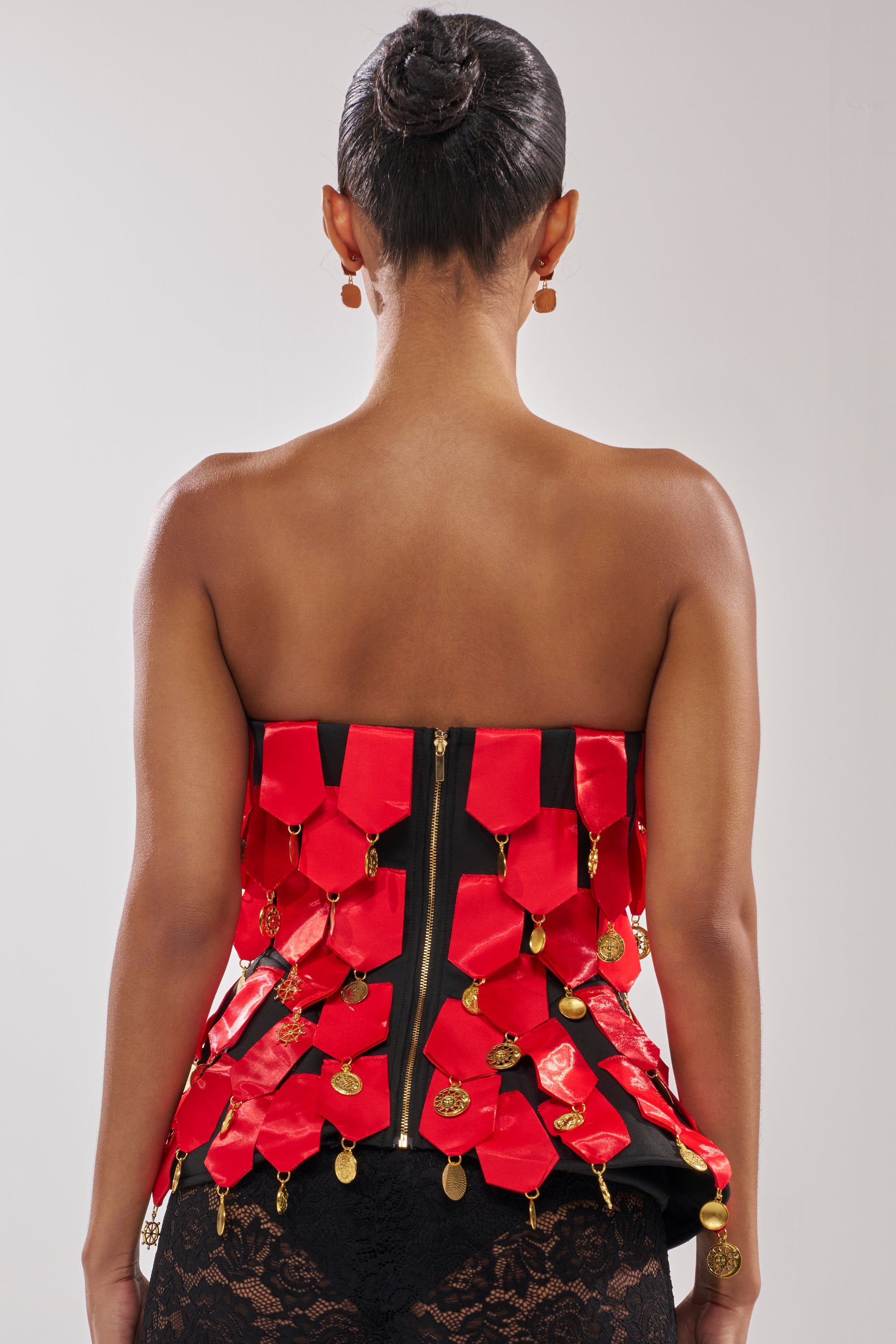 A woman with her back to the camera models the NOBODY KNOWS CORSET, a black top featuring red satin ribbons and gold charms, styled with black lace bottoms. Her hair is in a neat bun and she wears round earrings.