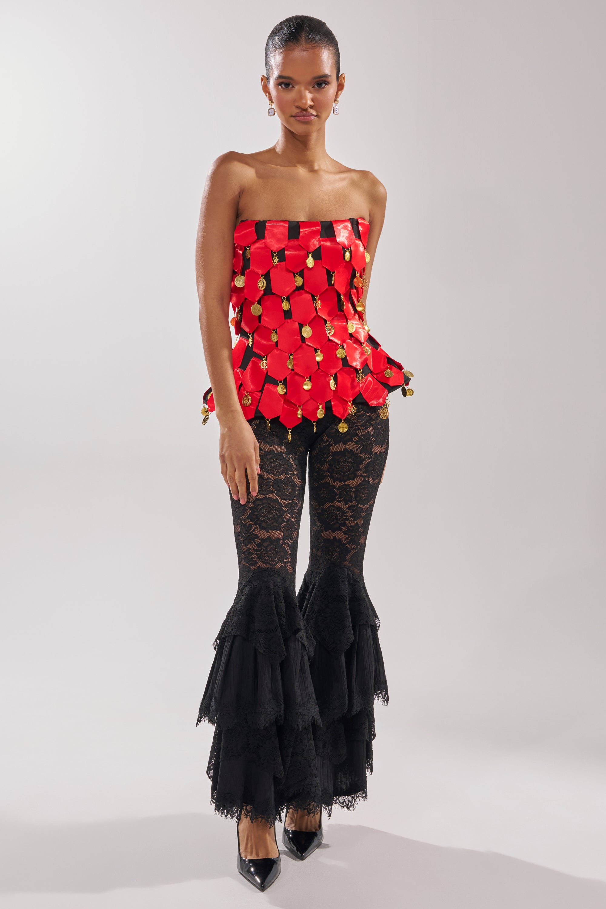 A woman stands against a plain background wearing the NOBODY KNOWS CORSET, a red satin tile-patterned top with gold accents, paired with black lace flared pants featuring layered ruffles and black pointed heels.