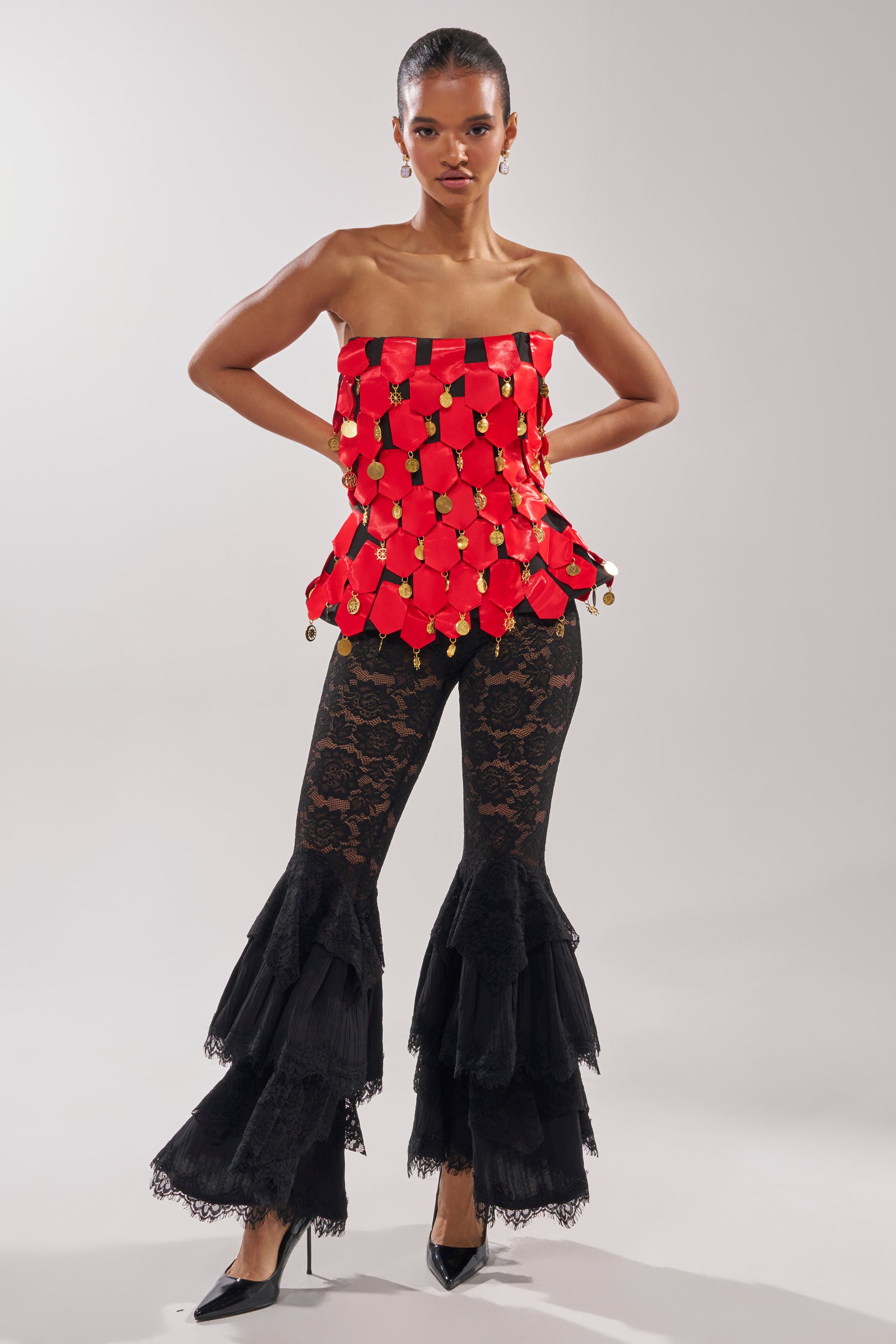 A woman poses in the NOBODY KNOWS CORSET, a strapless red satin top with gold accents, paired with black lace flared pants with tiered ruffle hems and black pointed heels. The background is a plain light gray.