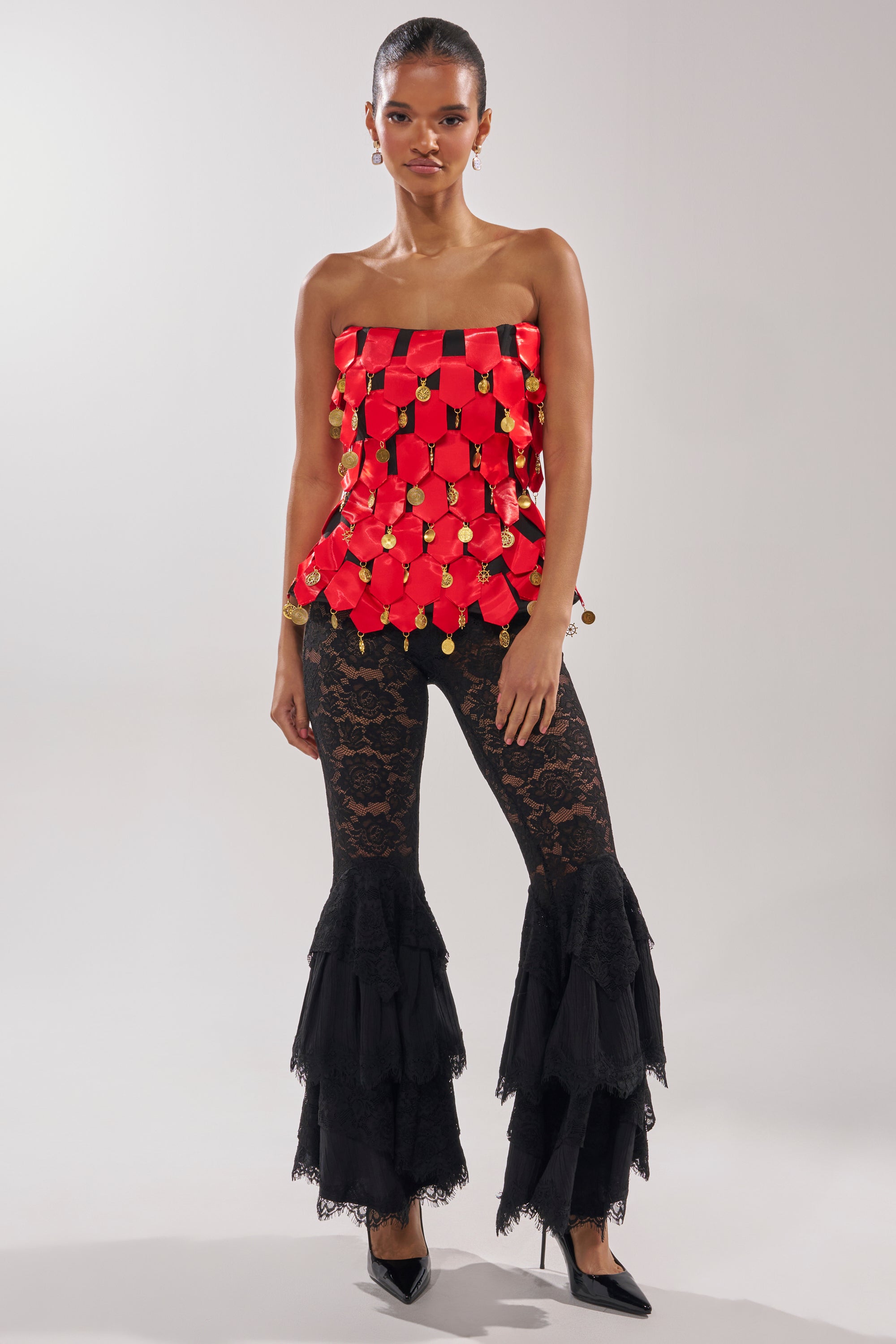 A woman wears the NOBODY KNOWS CORSET, a strapless red satin piece with gold details, paired with layered black lace flared pants and black pointed heels. Her hair is pulled back against a plain light background.