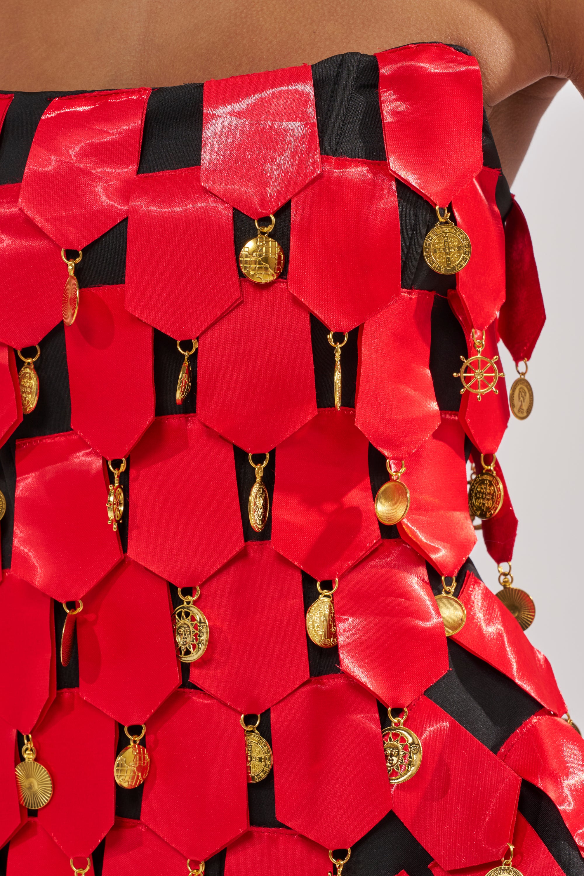 A close-up of the NOBODY KNOWS CORSET, crafted from shiny red satin panels joined together, each adorned with dangling gold charms, set against a black background.
