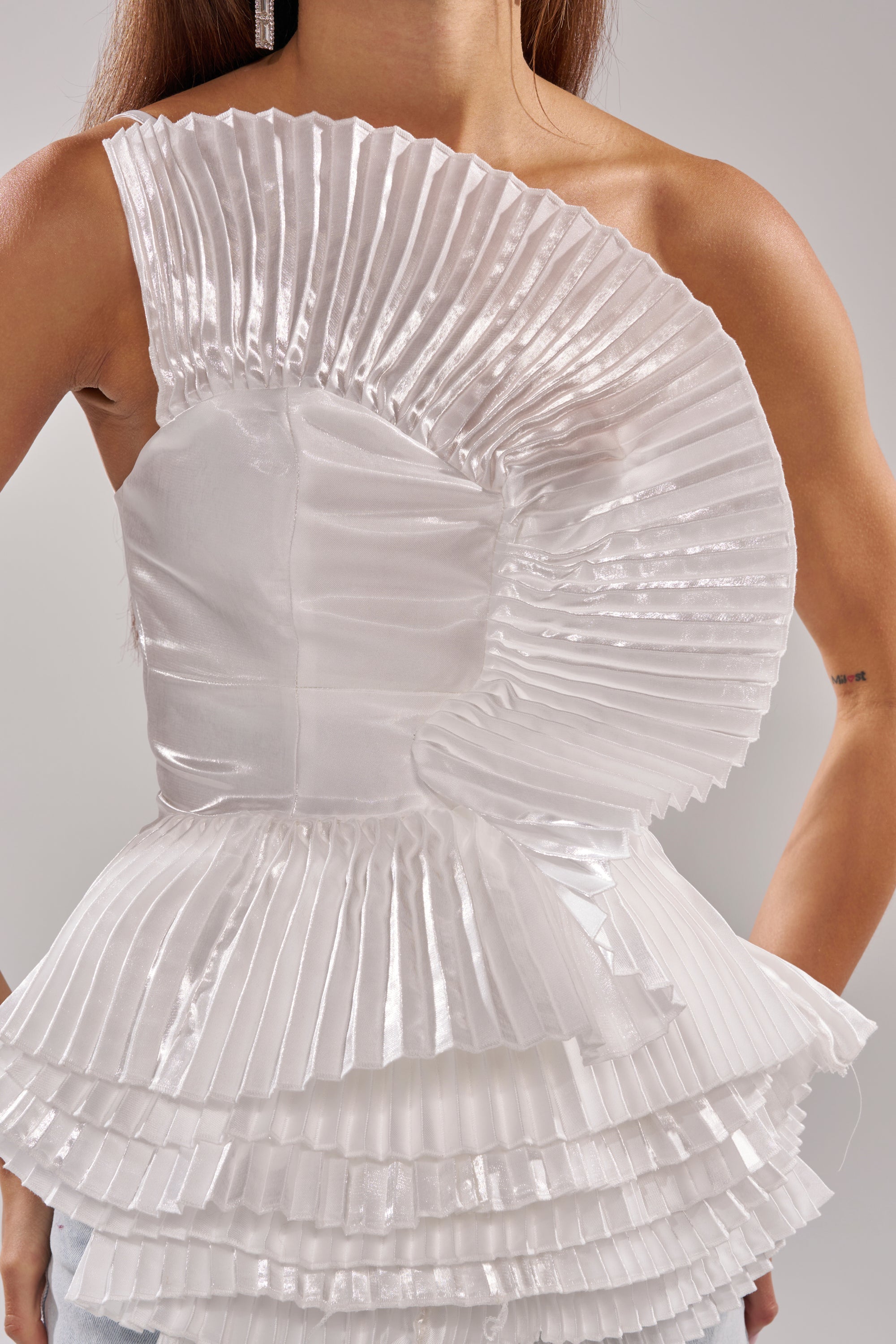 A person wears a shiny, white SUNSHINE PLEATED RUFFLE BLOUSE featuring asymmetrical, layered ruffles with a fan-like sculptural design accenting one shoulder and the waist. Only their upper body and arms are visible. 9