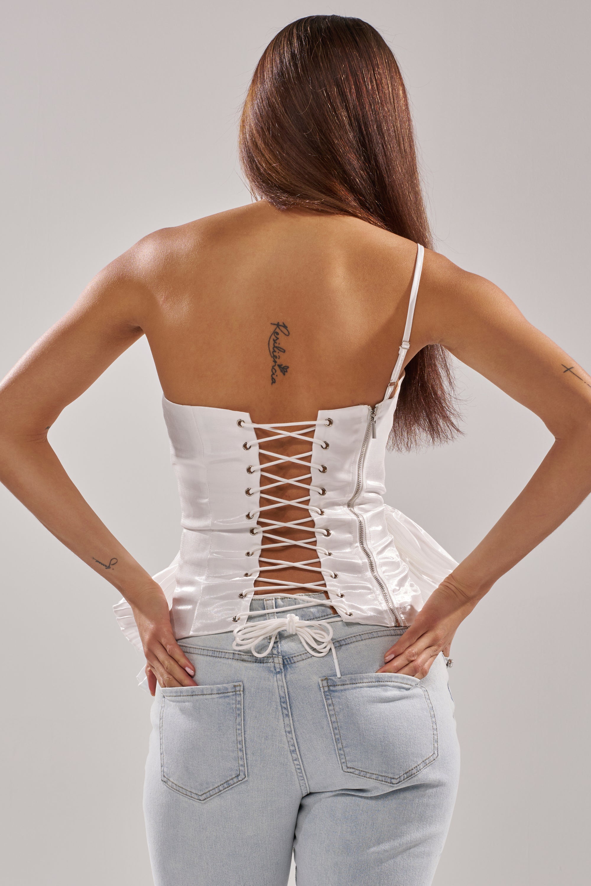 A woman with long straight brown hair, seen from behind, wears the SUNSHINE PLEATED RUFFLE BLOUSE and light blue jeans. She has upper back and left forearm tattoos and rests her hands on her hips. 5