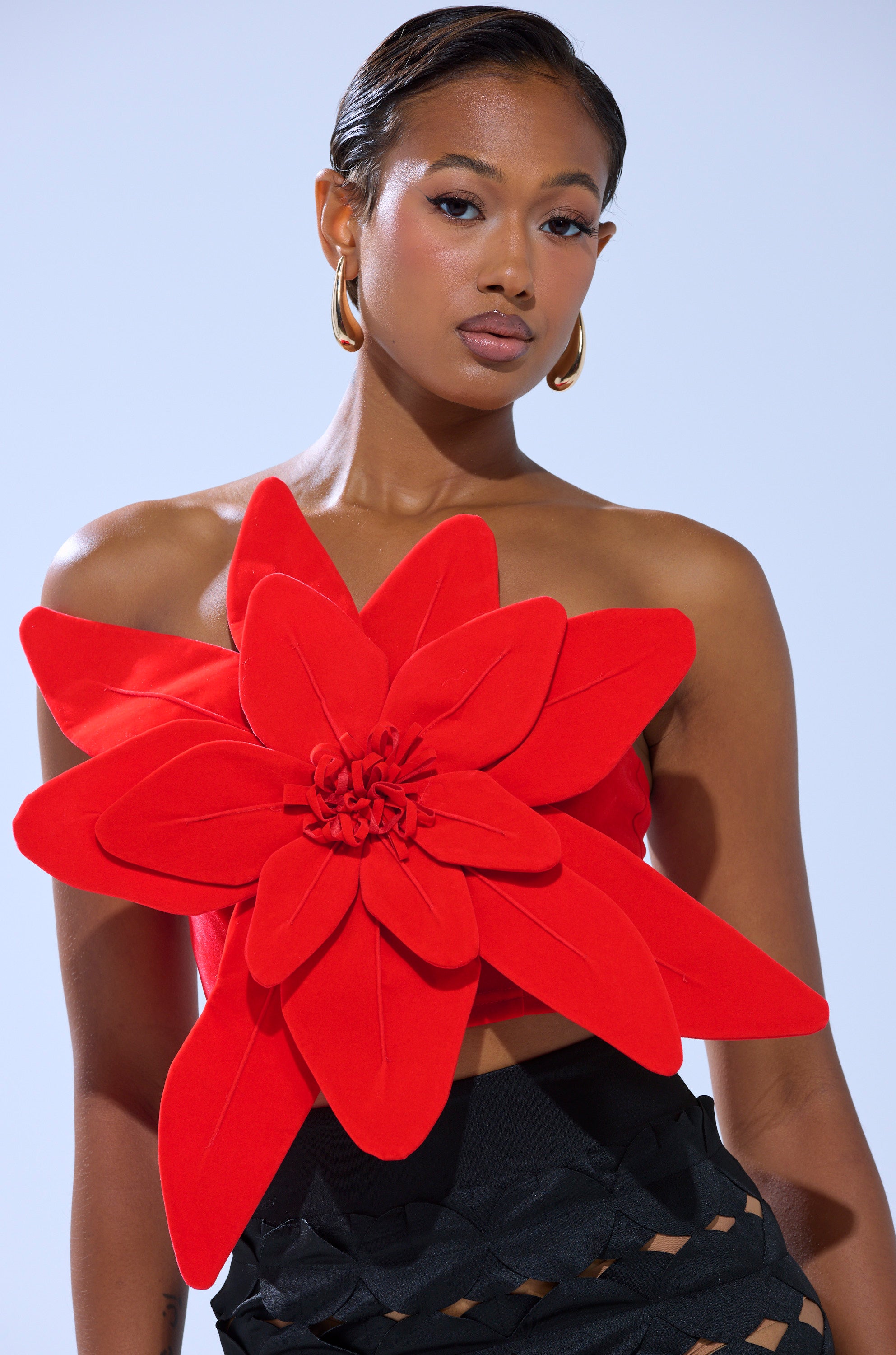 A woman with slicked-back hair and gold hoop earrings wears the HELLA MELODIES FLOWER TOP, a striking red velvet piece shaped like oversized petals, paired with black high-waisted pants featuring cut-out details.