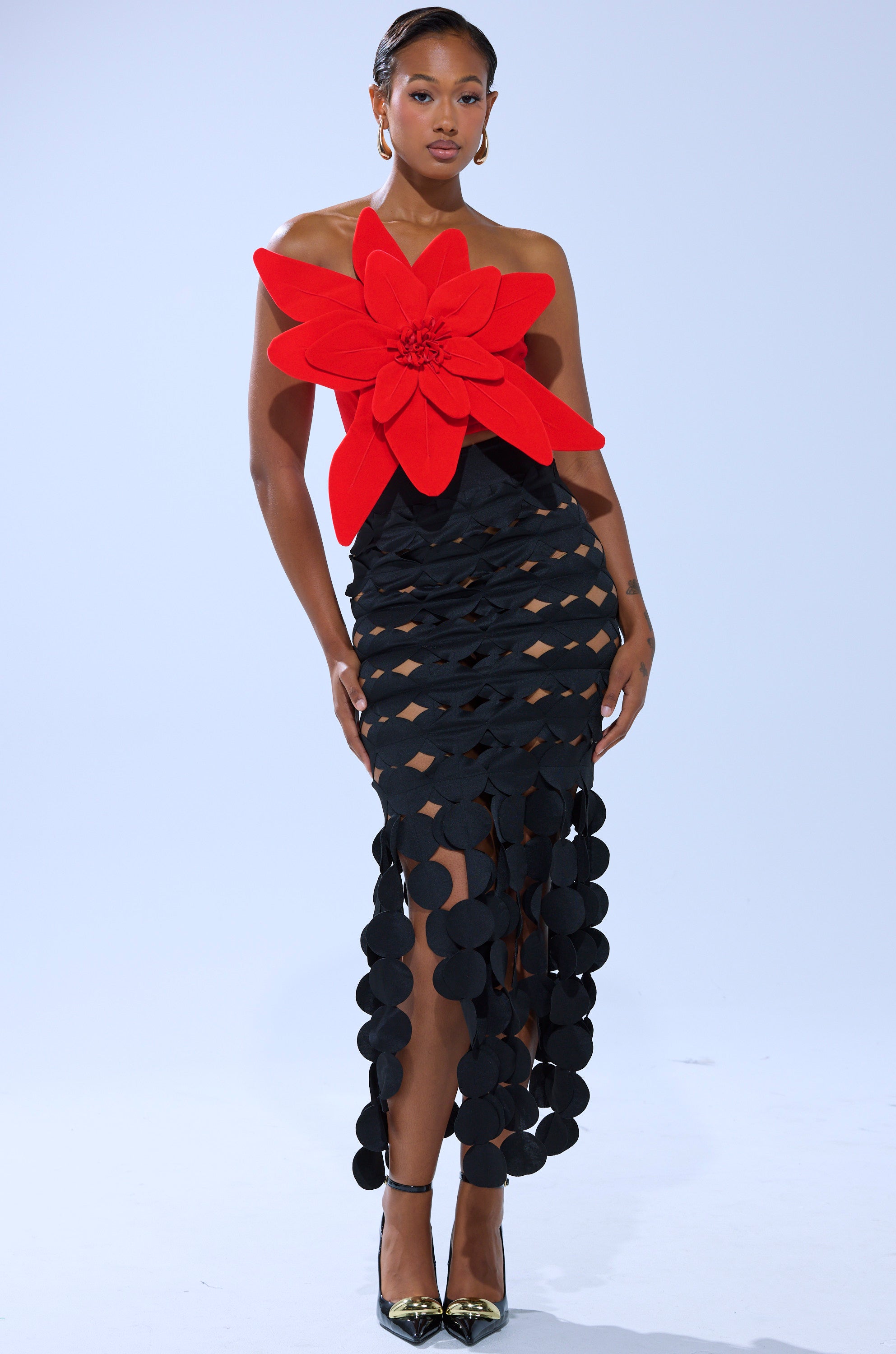A woman models the HELLA MELODIES FLOWER TOP—red velvet with an oversized flower applique on one shoulder—paired with a black circle-cut skirt and gold pointed-toe heels, set against a plain light background.